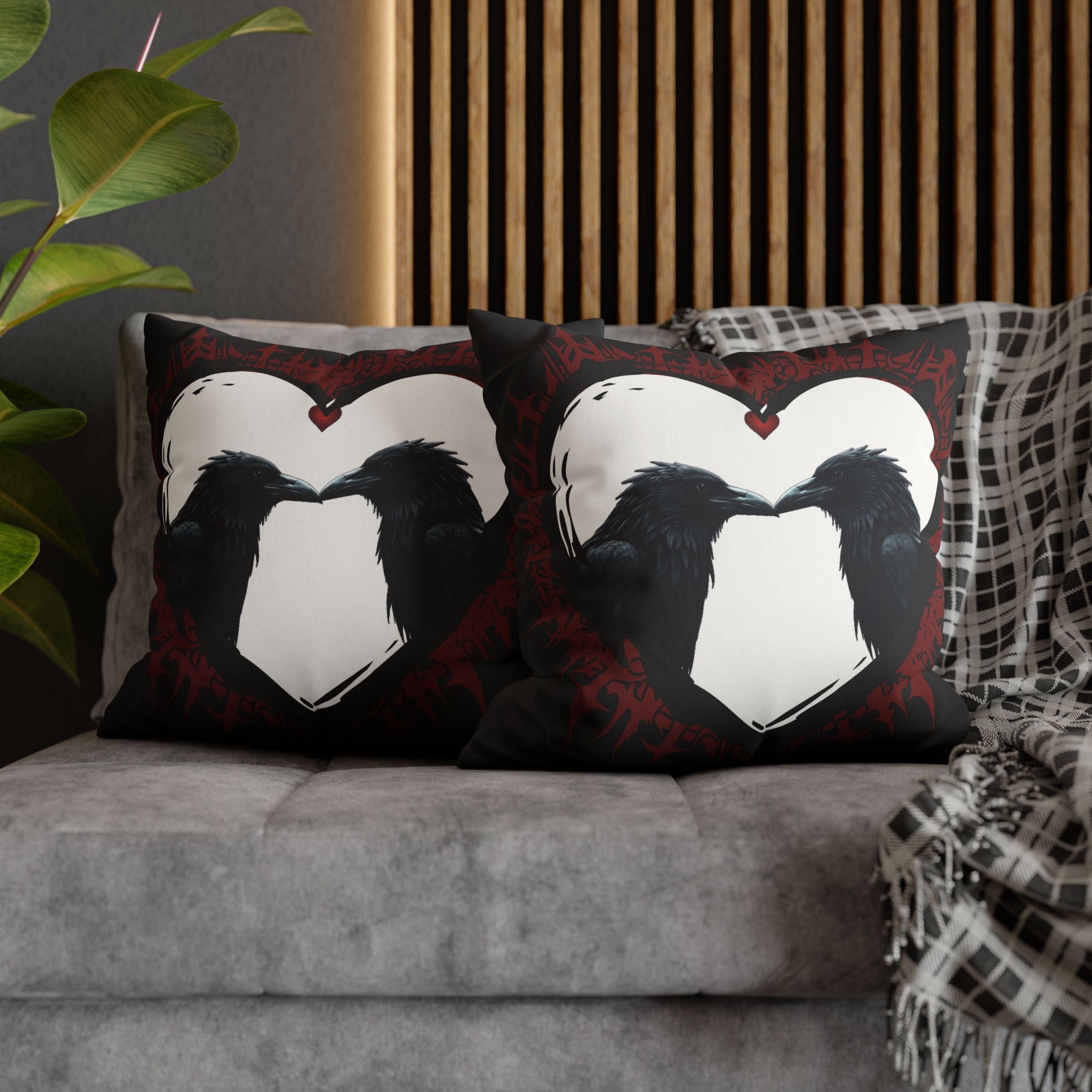 Two square throw pillows on a grey sofa featuring two ravens facing each other with beaks nearly touching inside a large white heart with a small red heart pendant at the top against a black background with deep crimson gothic vine and tribal details radiating outward