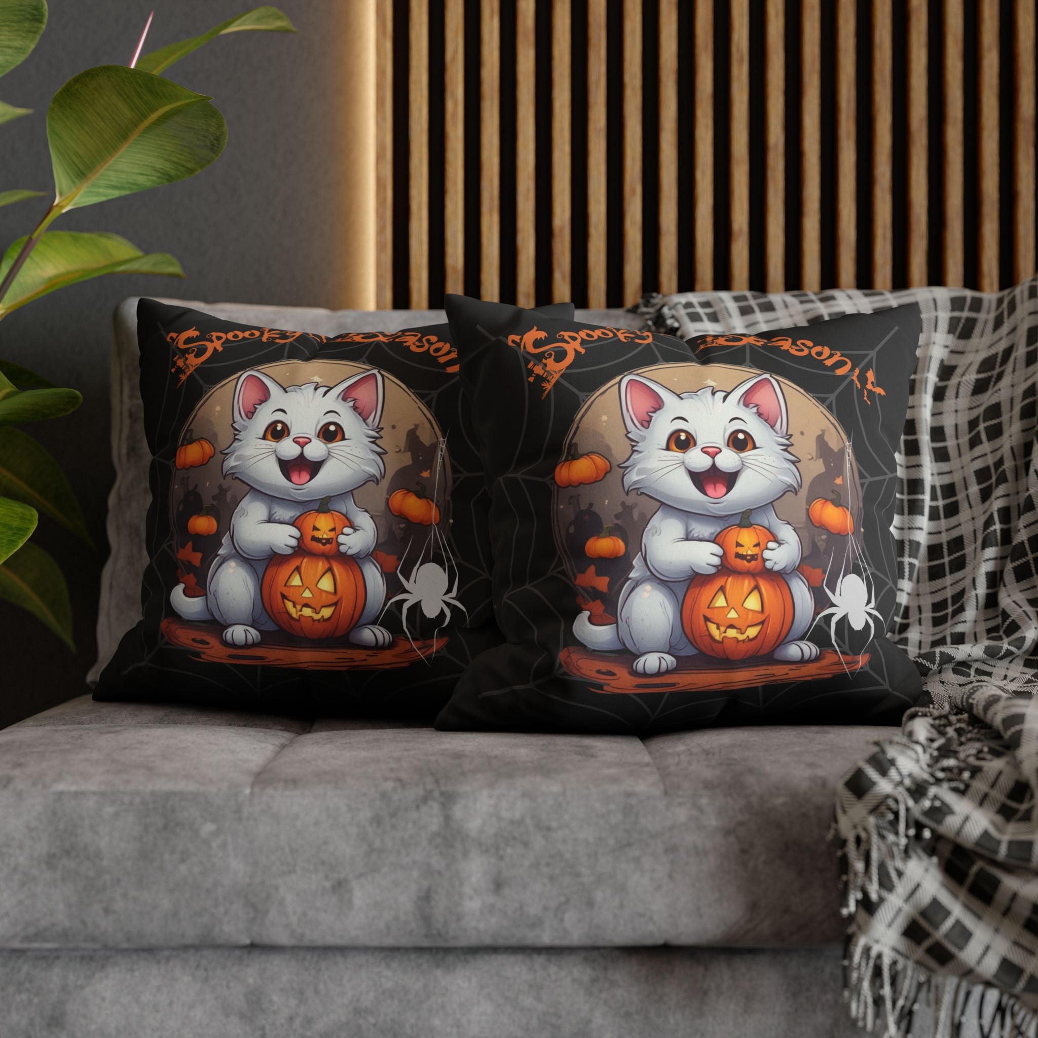 Two square throw pillows on a grey sofa featuring a grinning white cat clutching a glowing jack-o'-lantern beneath a full moon with floating mini pumpkins, a dangling white spider, and Spooky Season orange script over a grey cobweb black background