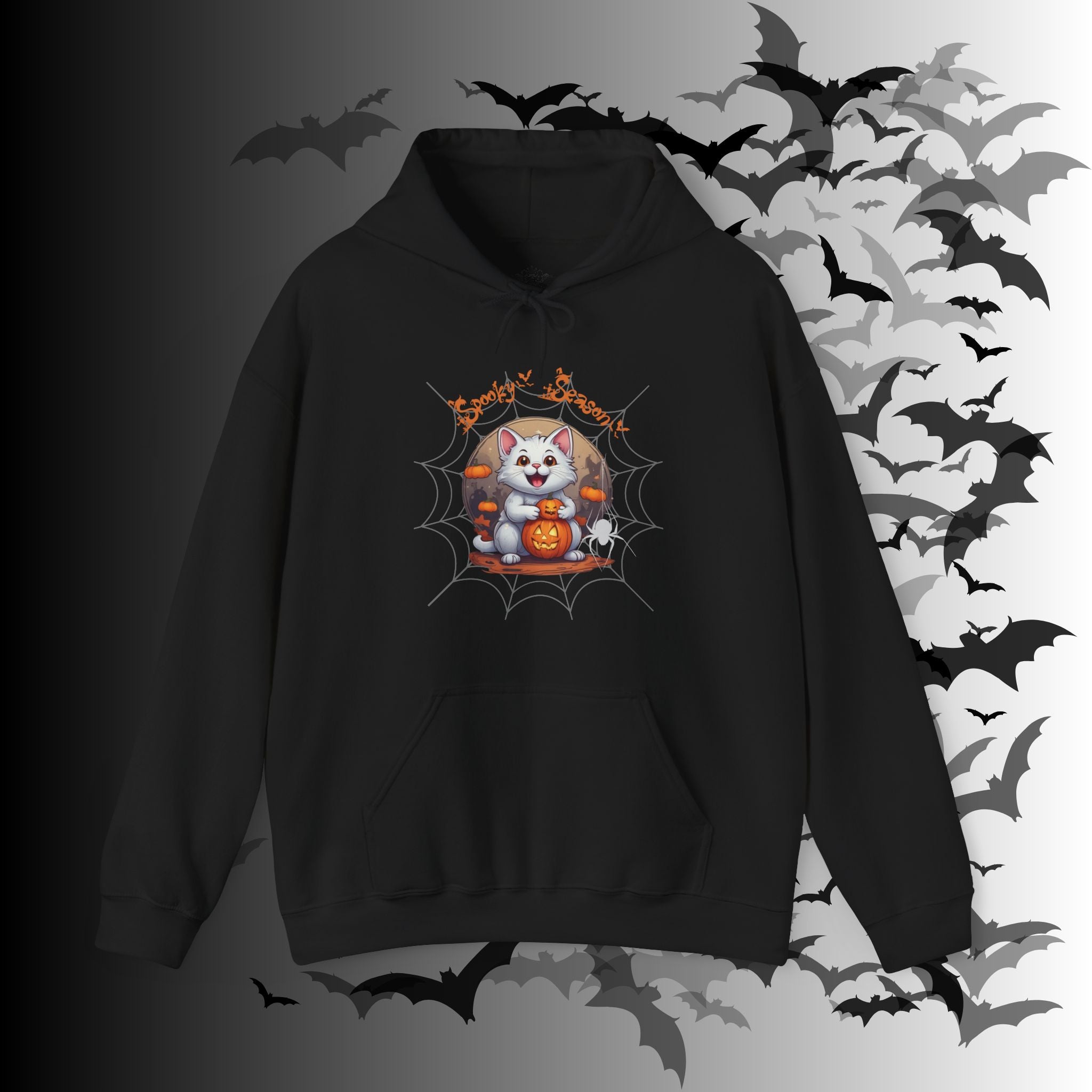 Black pullover hoodie with kawaii white cat holding a jack-o-lantern inside a spider web frame with "Spooky Season" text in orange gothic lettering on a bat-filled background