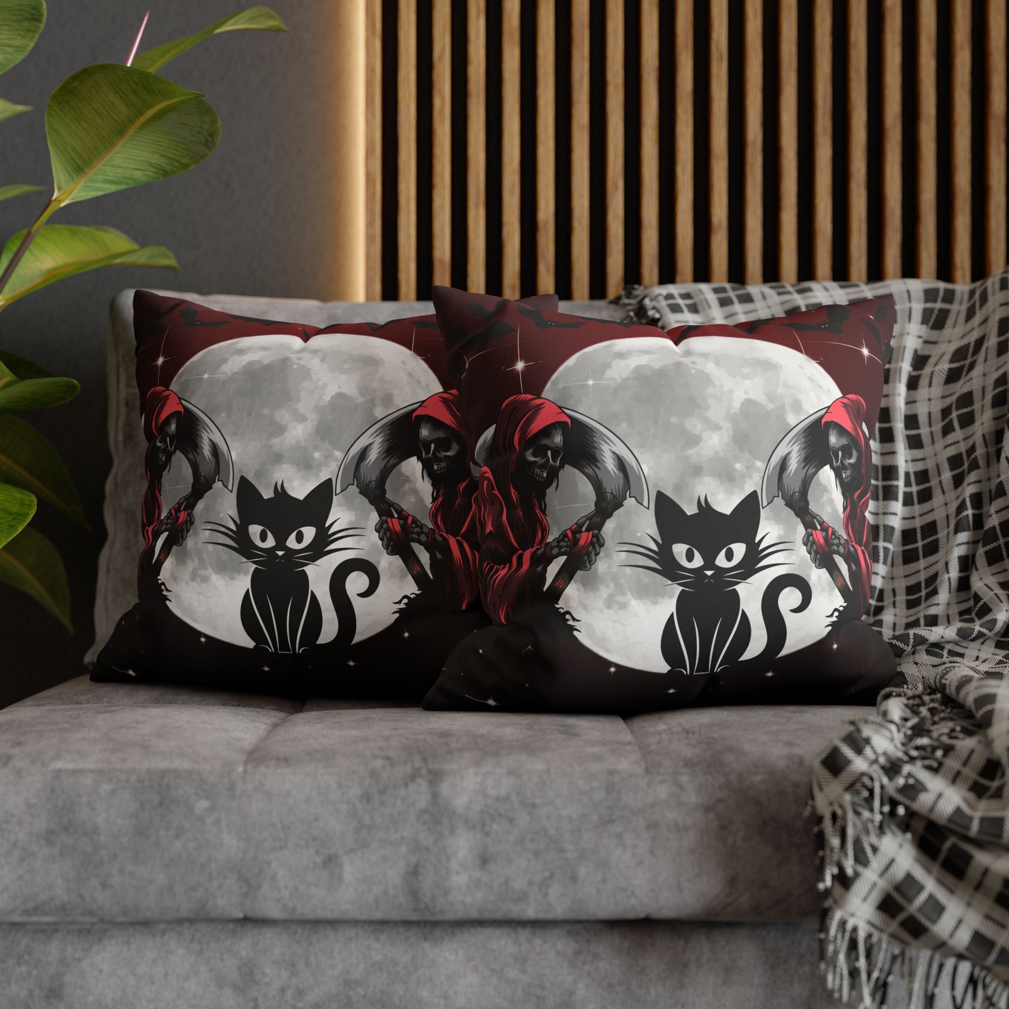 Two square throw pillows on a grey sofa featuring a bold black cat silhouette with sharp eyes seated before a glowing full moon with a red-cloaked skeletal Grim Reaper holding a scythe with dark wings spread against a deep crimson and black starfield background