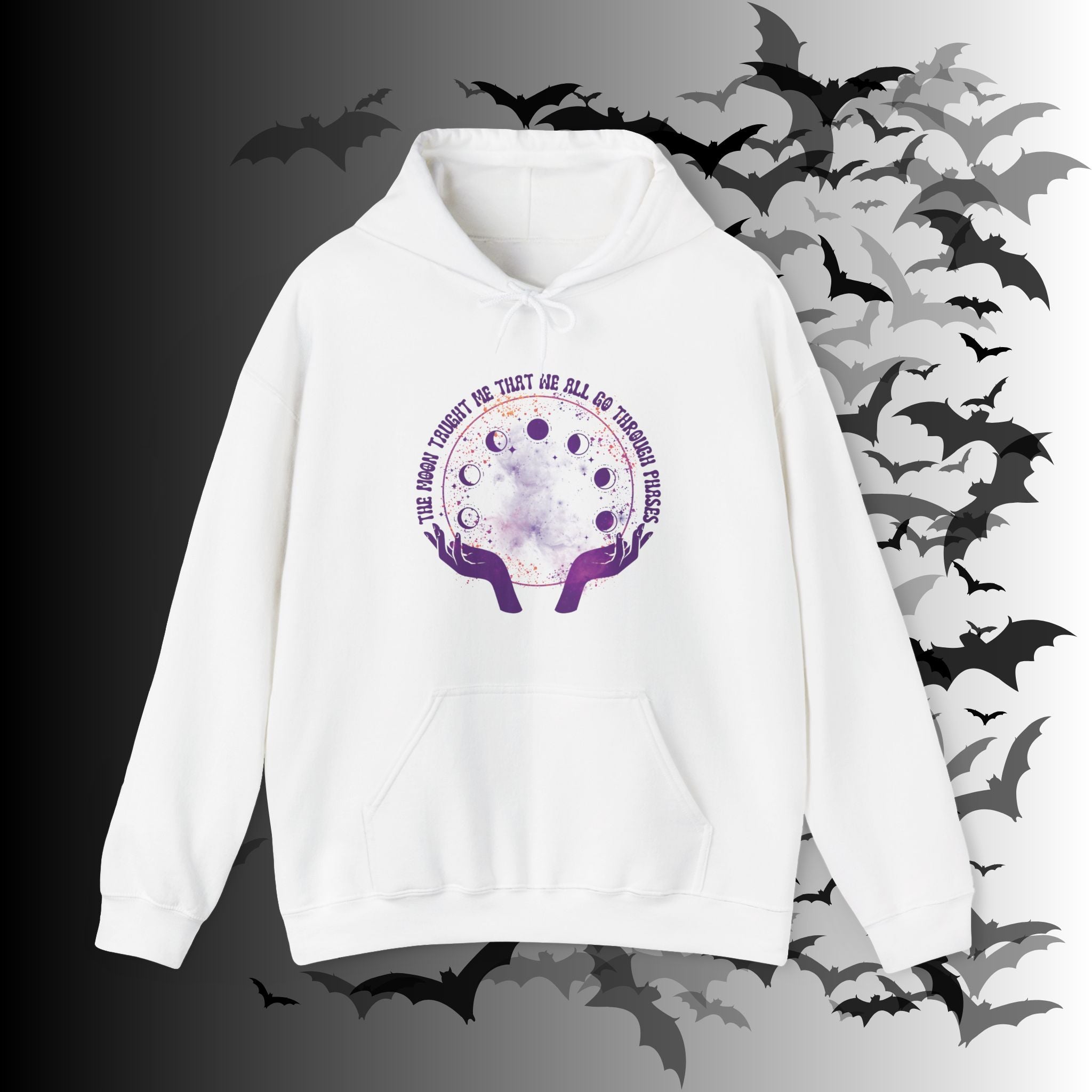 White pullover hoodie with purple witch hands cradling a watercolor galaxy orb showing moon phases with "The Moon Taught Me That We All Go Through Phases" script circling the top on a bat-filled background