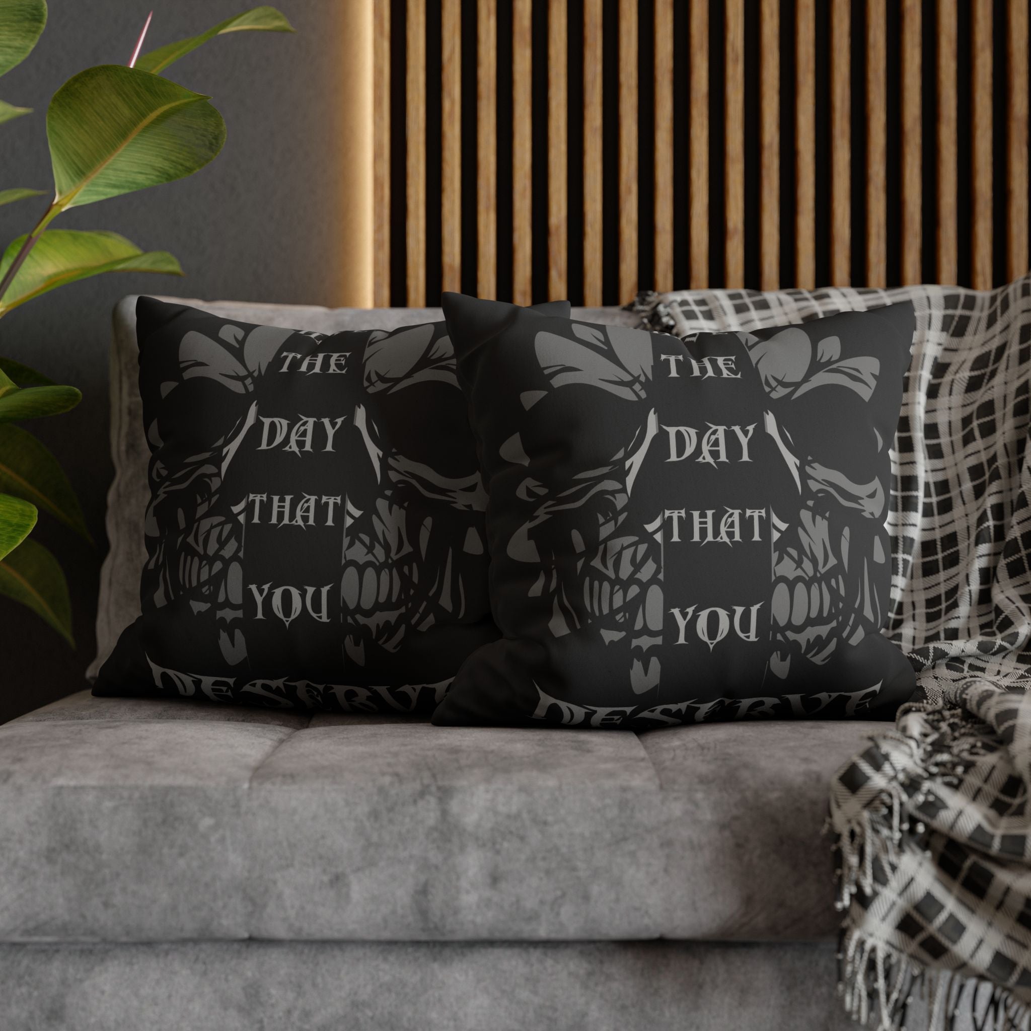 Two square throw pillows on a grey sofa featuring a snarling skull centered within a large dark floral and bow motif with gothic blackletter text reading The Day That You Die rendered in deep charcoal grey on black