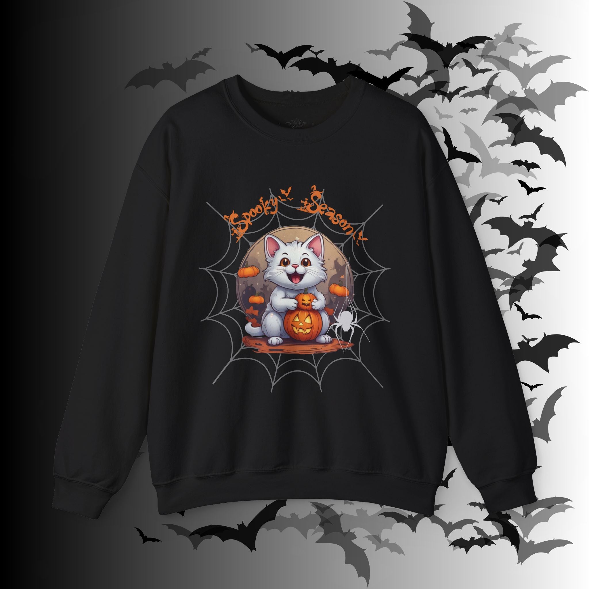 Black crewneck sweatshirt with kawaii white cat holding a jack-o-lantern inside a spider web frame with "Spooky Season" text in orange gothic lettering on a bat-filled background