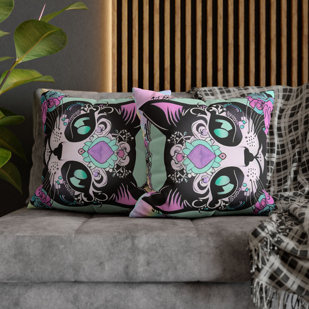 Two square throw pillows on a grey sofa featuring a close-cropped sugar skull cat face with teal gem eyes, intricate black filigree, a faceted purple diamond jewel at center, and hot pink roses against a holographic pastel mint and lavender background