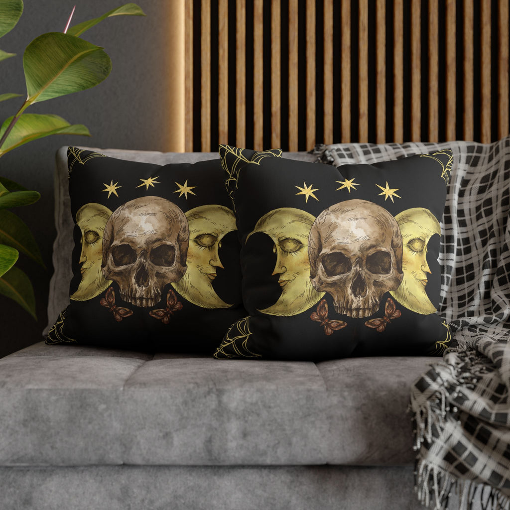 Two square throw pillows on a grey sofa featuring a weathered skull flanked by two golden crescent moon faces with closed eyes, crowned by three eight-point gold stars with small brown butterflies below and gold line details at the corners against a deep black background