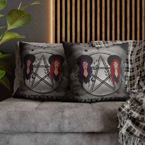Two square throw pillows on a grey sofa featuring two illustrated witches in wide-brimmed black hats with purple and red hair flanking a hand-drawn pentagram wrapped in botanical branches with Best Witches gothic script above and below on a smoky grey background