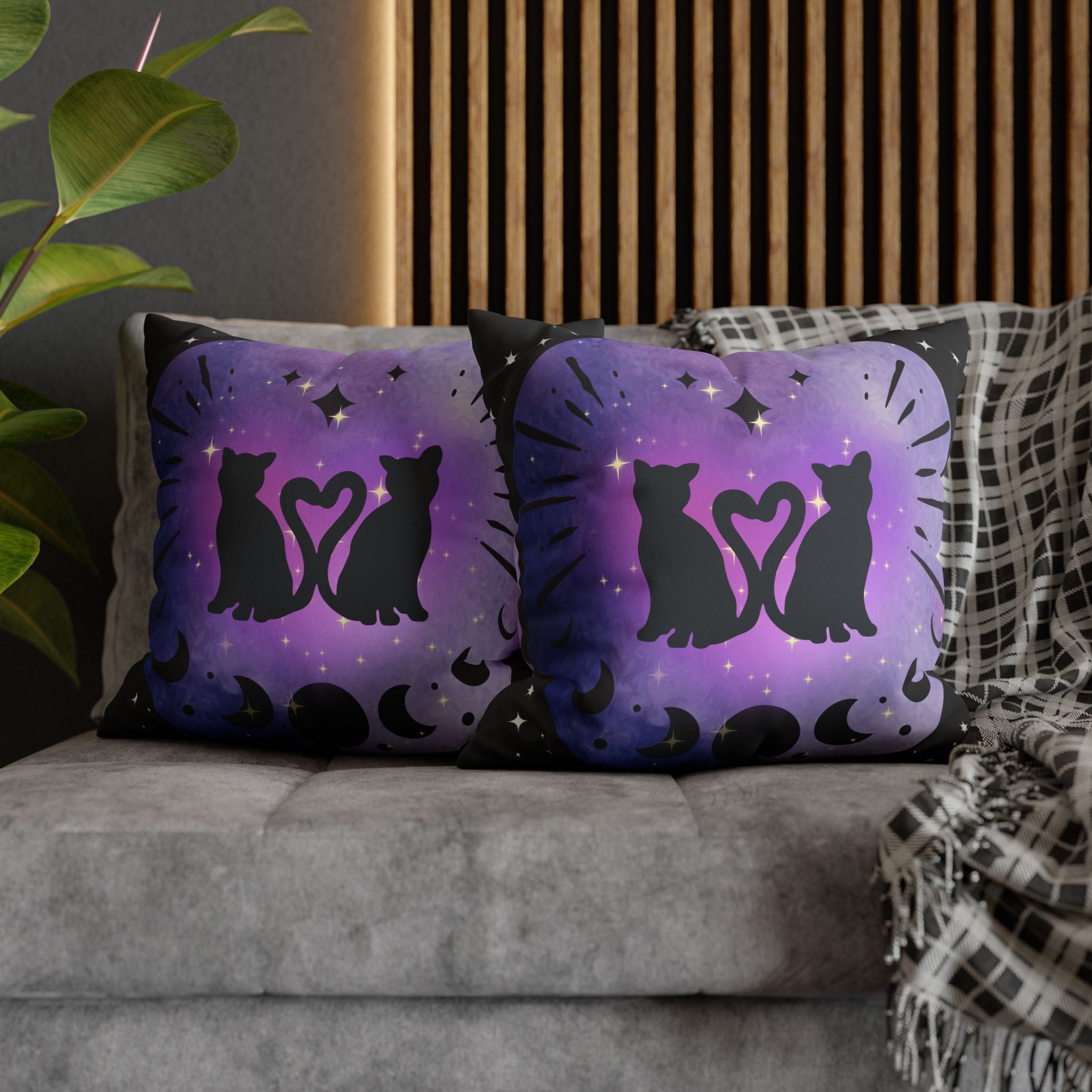 Two square throw pillows on a grey sofa featuring two black cat silhouettes facing each other with tails curled into a heart shape inside a soft purple to indigo watercolor vignette with moon phase sequence along the bottom and gold diamond stars on deep black