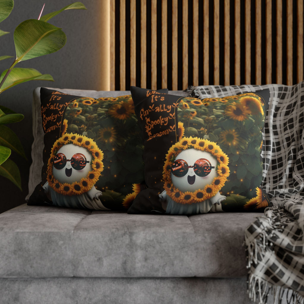 Two square throw pillows on a grey sofa featuring a round ghost wearing a sunflower crown and jack-o'-lantern sunglasses floating before a lush glowing sunflower field with orange script reading When It's Finally Spooky Season against a deep black background