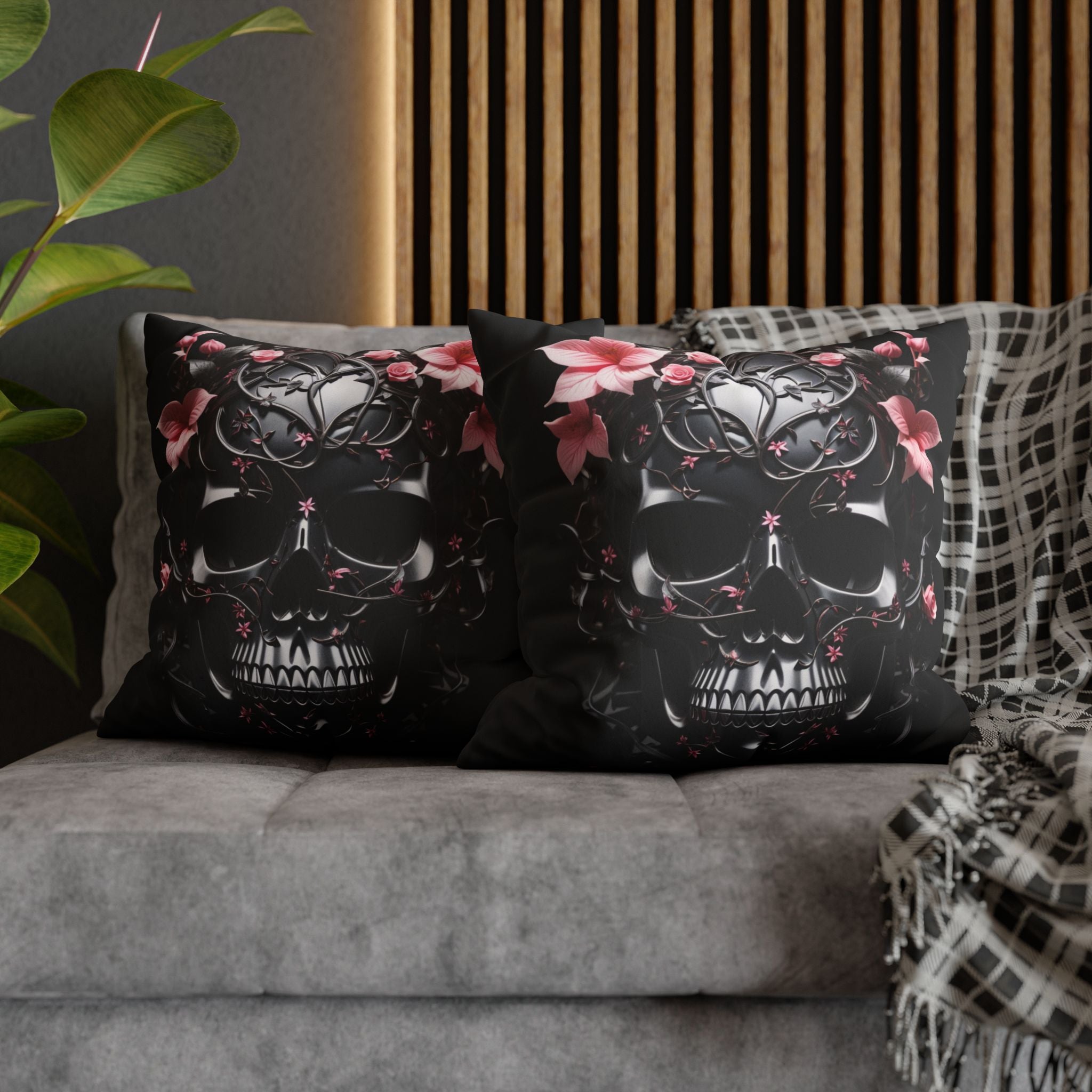 Two square throw pillows on a grey sofa featuring a large obsidian skull covered in intricate black iron vine and filigree work with soft pink roses and lilies blooming from the crown and eye sockets against a deep black background