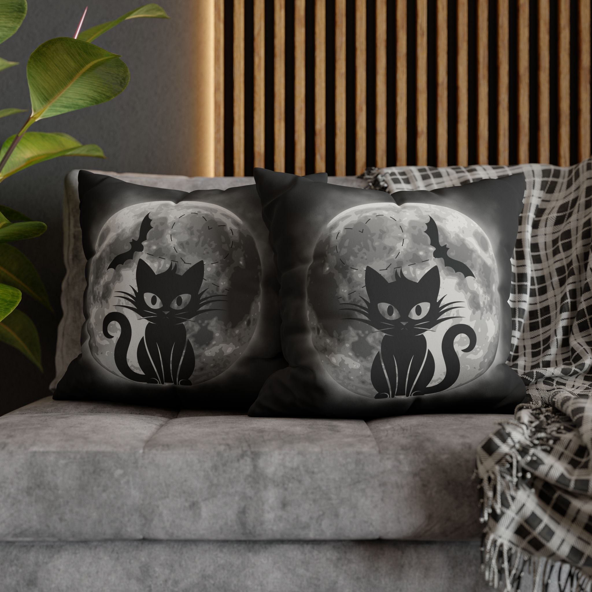 Two square throw pillows on a grey sofa featuring a black cat silhouette with curled tail seated before a large luminous full moon with a bat in flight overhead fading into deep black mist