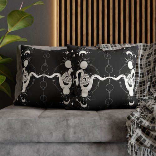 Two square throw pillows on a grey sofa featuring a pale coiling serpent with a sun face etched into its body surrounded by crescent moons, geometric orbital rings, starburst sparkles, and delicate constellation lines in white linework on a deep black background