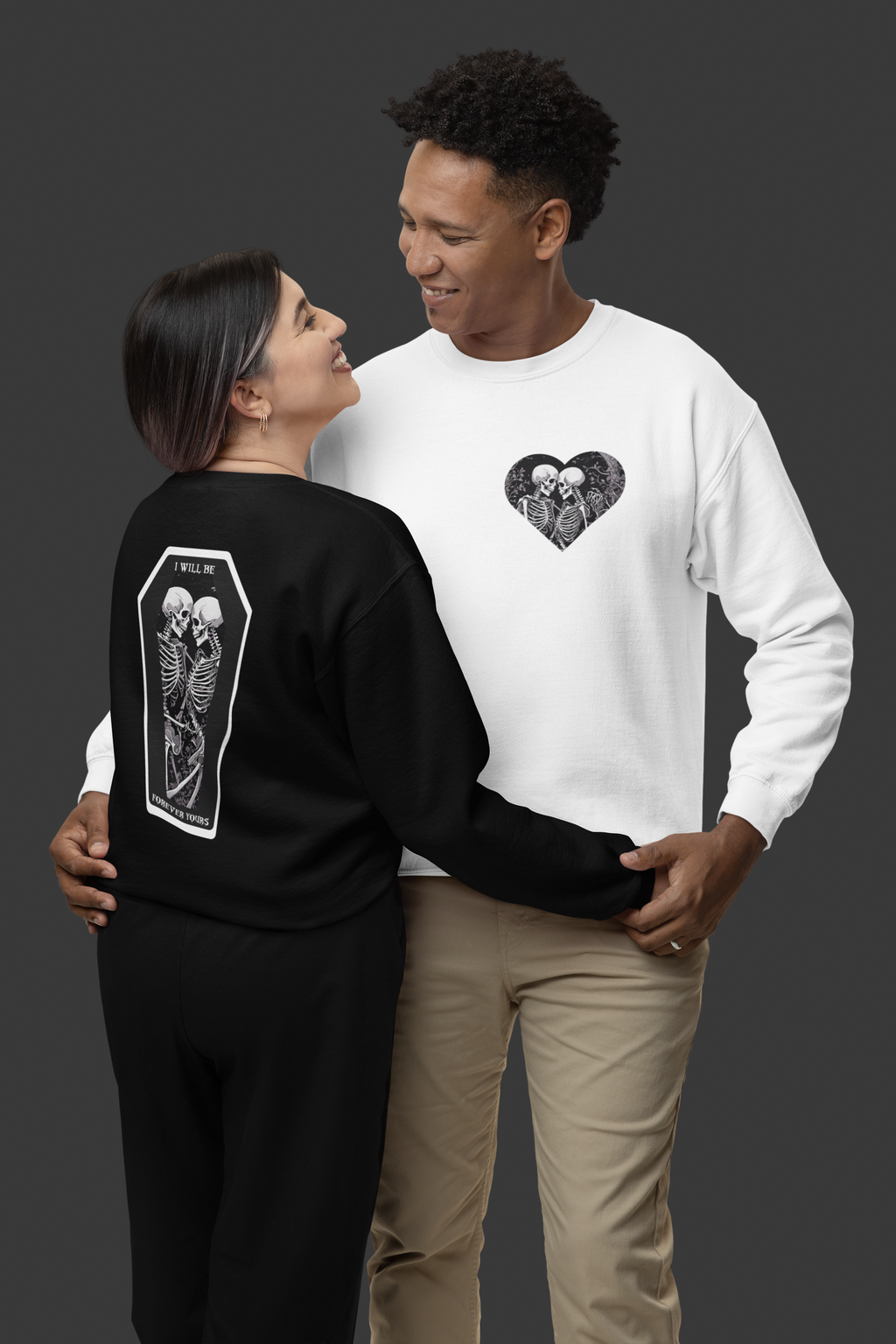Gothic crewneck sweatshirt shown in black displaying back coffin print with embracing skeletons and "I Will Be Forever Yours" text, and white displaying front chest skeleton heart print, worn by a couple