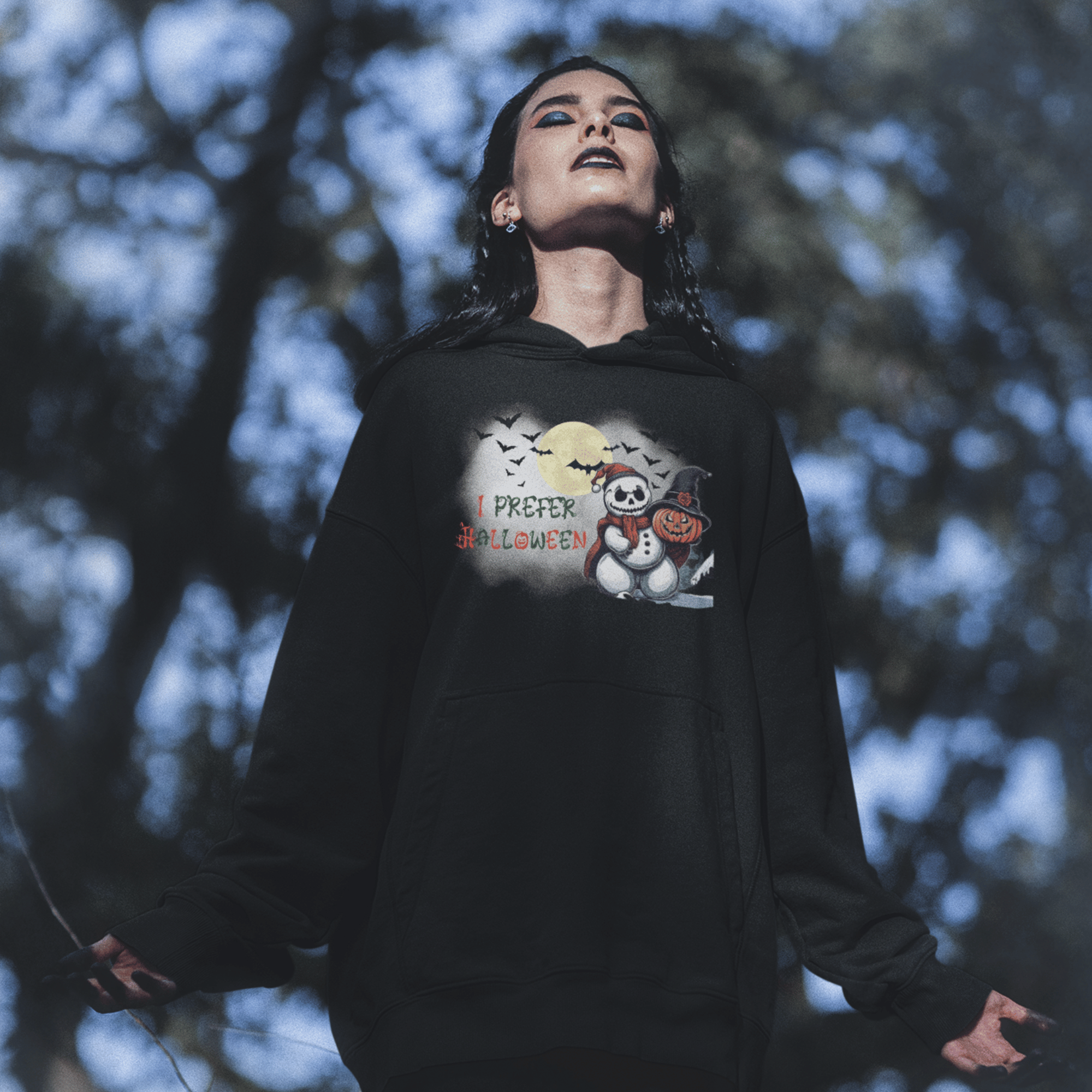 Black hoodie with skull-faced snowman in jester hat, jack-o-lantern in witch hat, bats, full moon, and "I Prefer Halloween" text worn by a woman outdoors