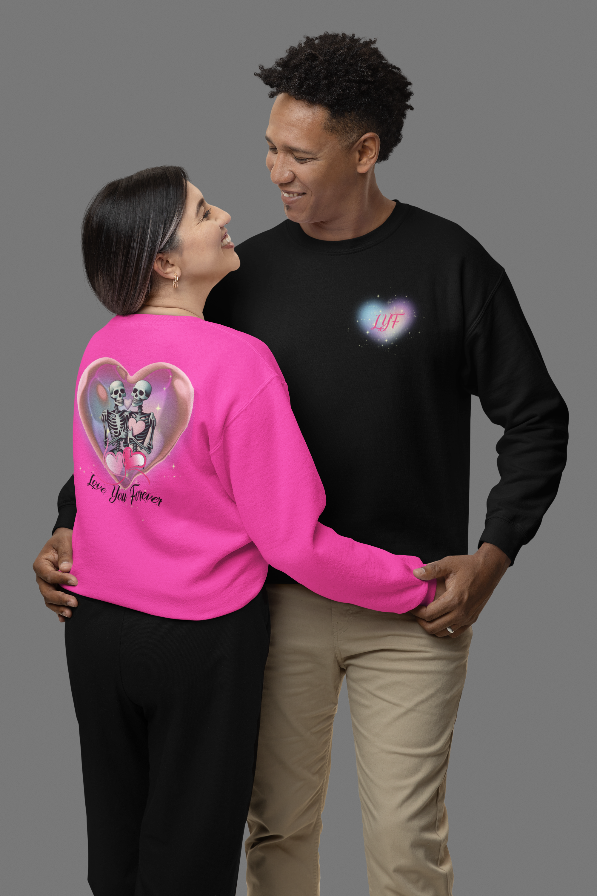 Gothic Valentine's crewneck sweatshirt shown in black with "LYF" chest print and heliconia pink with back print of two skeletons inside a glowing pink heart bubble with "Love You Forever" script worn by a couple in a studio