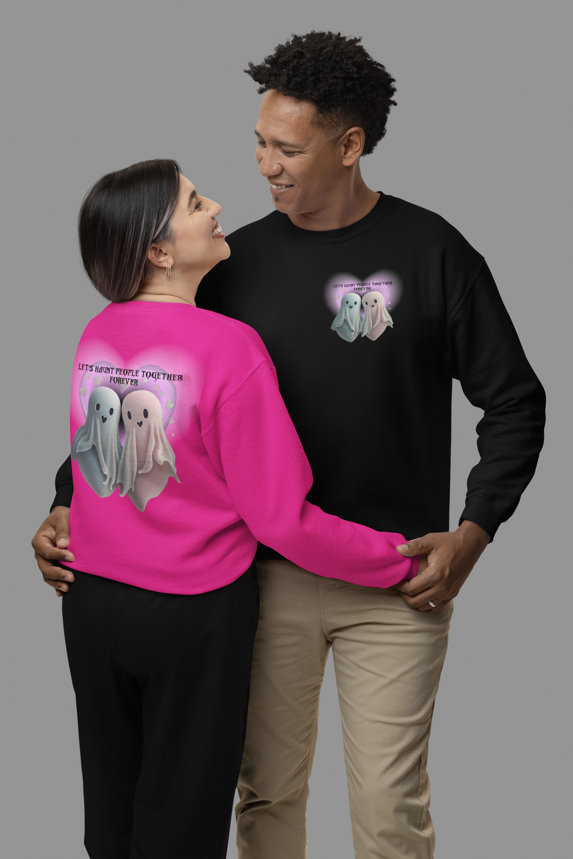 Gothic Valentine's crewneck sweatshirt shown in heliconia pink with large back print of two kawaii ghosts inside a glowing heart and "Let's Haunt People Together Forever" text, and black with matching front chest print, worn by a couple in a studio