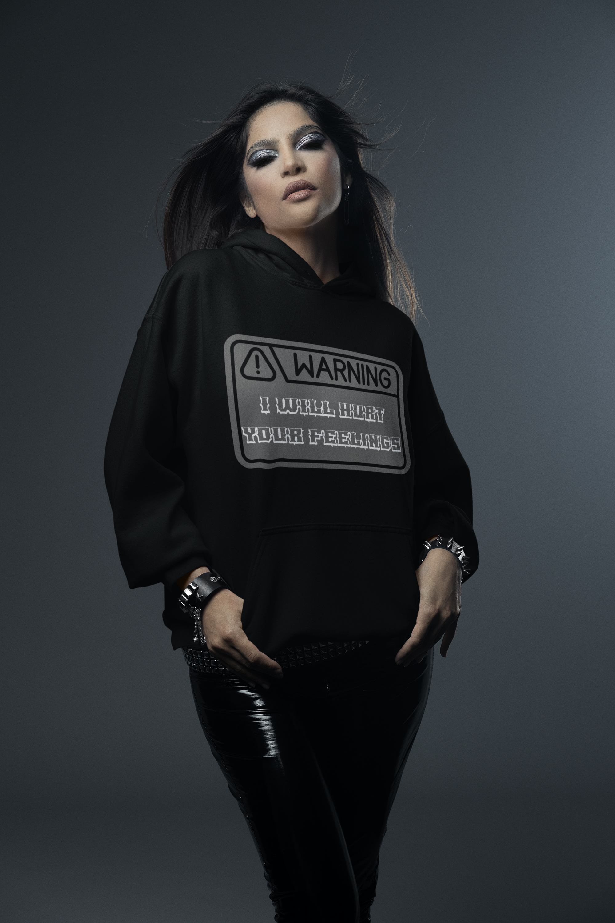Black pullover hoodie with grey warning label graphic and "WARNING — I Will Hurt Your Feelings" in blocky stencil lettering worn by a woman in gothic makeup and patent leather pants