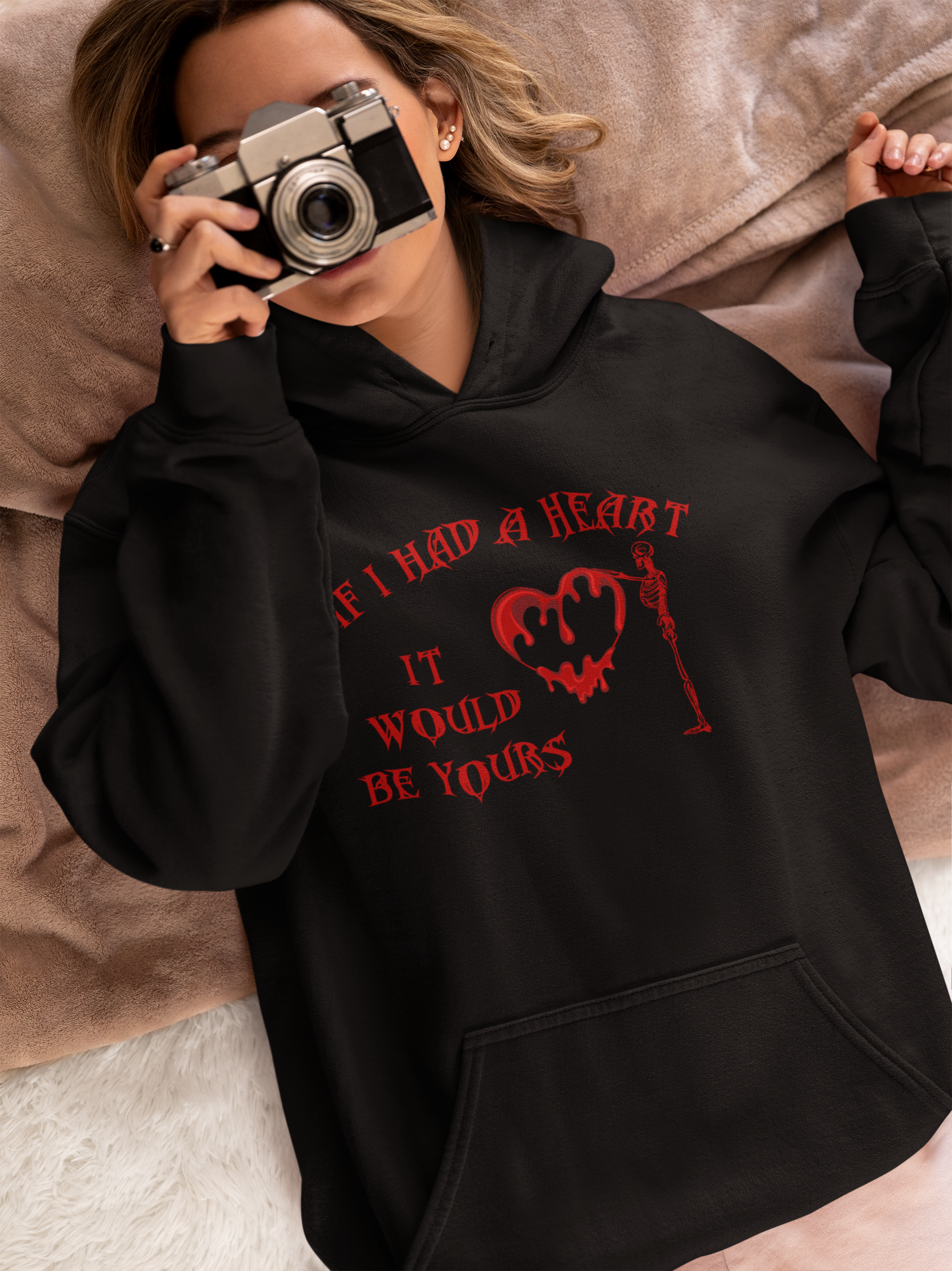 Black pullover hoodie with red gothic lettering "If I Had a Heart It Would Be Yours" with dripping melting heart and skeleton graphic worn by a woman lying in bed holding a vintage camera