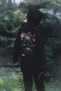 
Black pullover hoodie back print with a large ornate black skull covered in intricate vine detailing with pink cherry blossom flowers blooming across it worn by a woman in a forest
