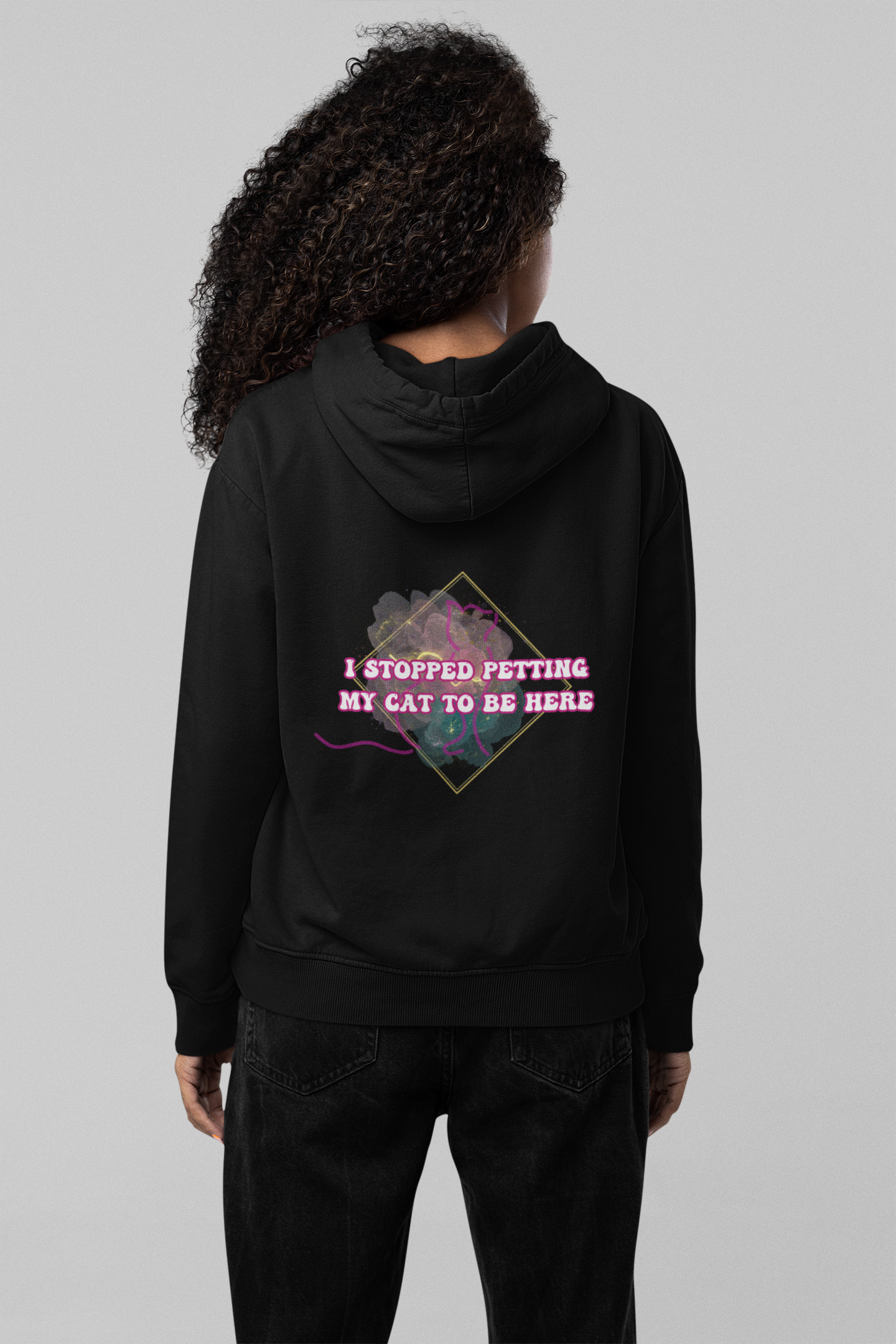 
Black pullover hoodie back print with "I Stopped Petting My Cat To Be Here" in pink retro lettering inside a gold diamond frame over a dark floral watercolor background worn by a woman with curly hair