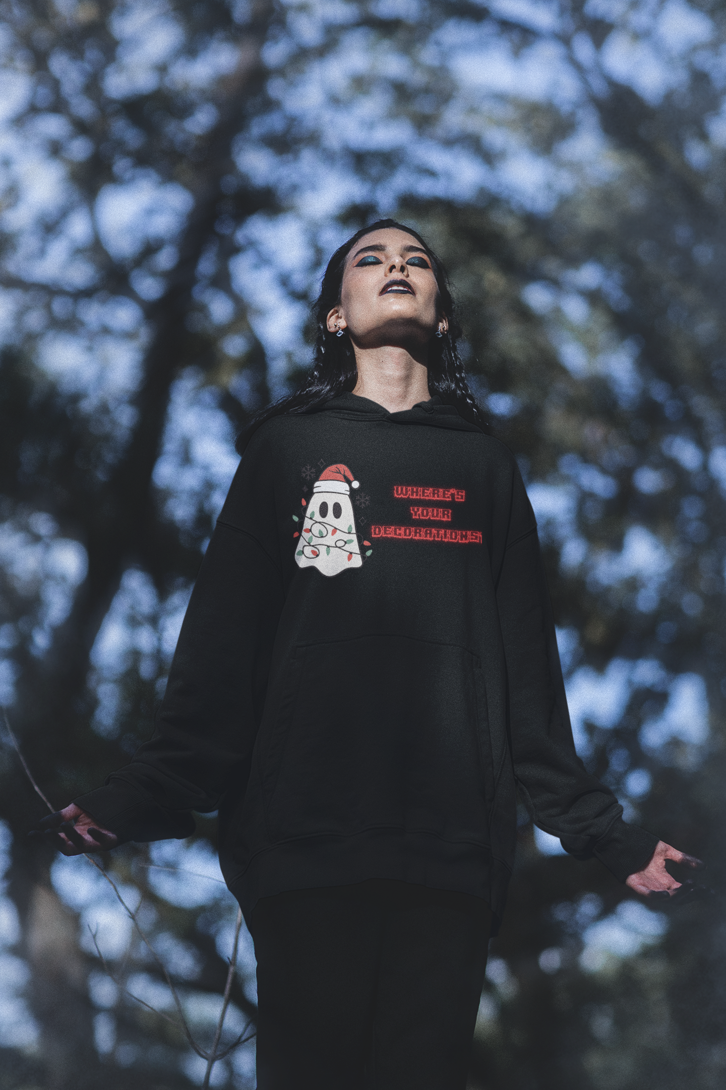 Black pullover hoodie with kawaii ghost in Santa hat wrapped in colorful Christmas lights and "Where's Your Decorations?" in red lettering worn by a woman in a forest