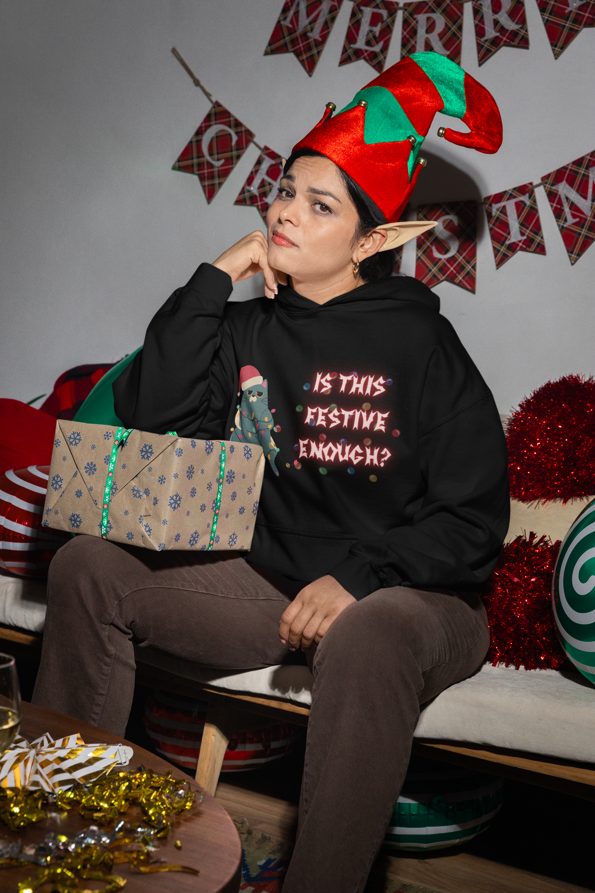 Black pullover hoodie with grumpy cat in Santa hat tangled in colorful Christmas lights and "Is This Festive Enough?" in glowing gothic lettering worn by a woman in an elf hat at a Christmas party