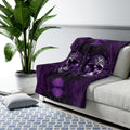 Dark purple and black throw blanket with large ornate skulls with glowing purple eyes against a Victorian damask floral background draped over a sofa