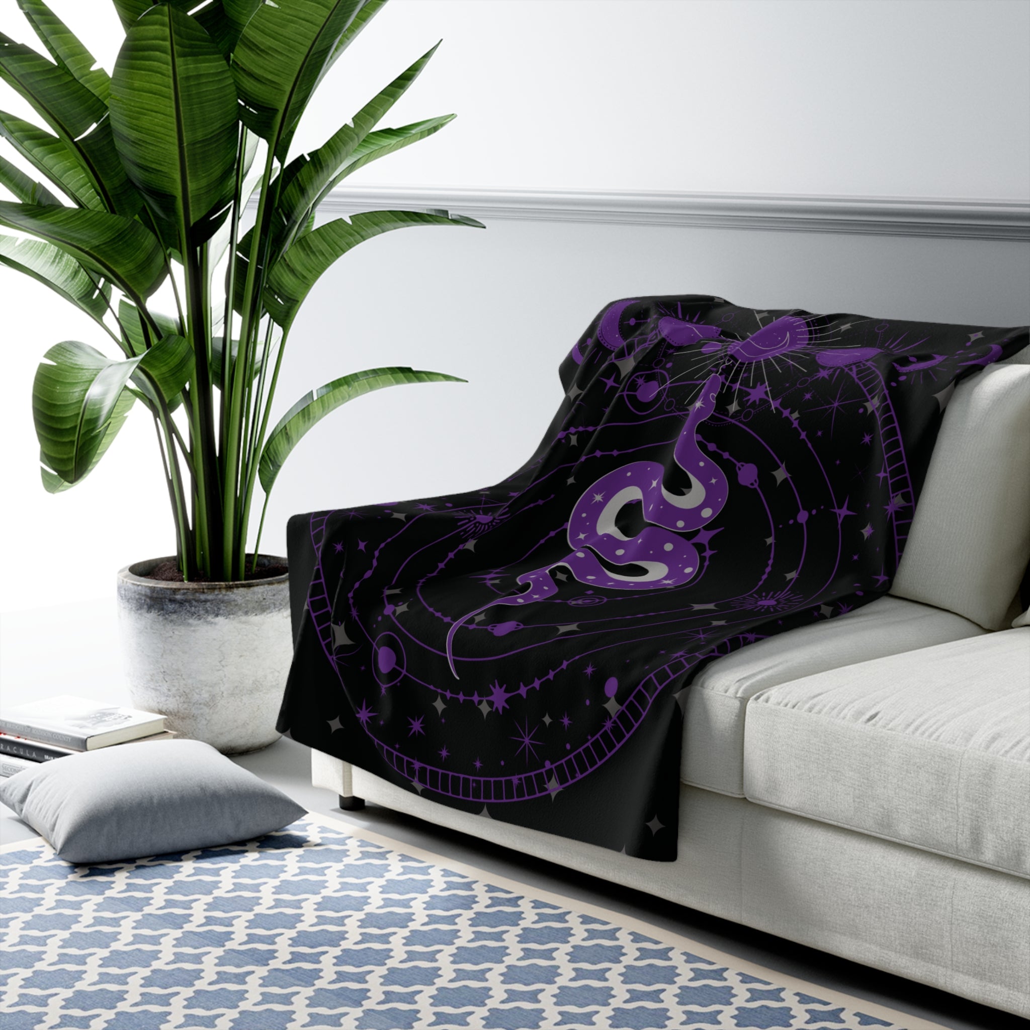 Black throw blanket with coiled purple snake surrounded by celestial star chart, constellations, orbiting rings, and all-seeing eye in dark grey linework draped over a sofa