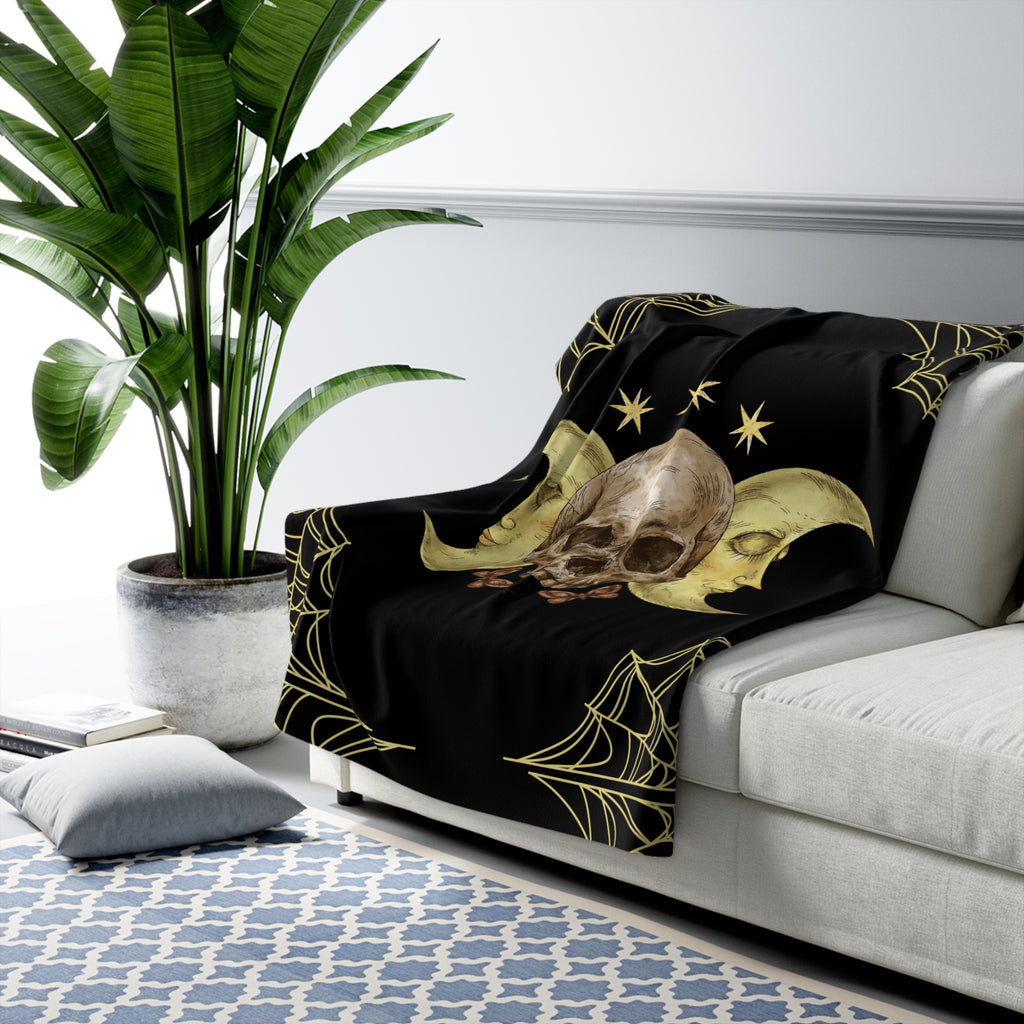 Black throw blanket with two sleeping gold crescent moon faces cradling a weathered skull with gold spider web corners and star accents draped over a sofa