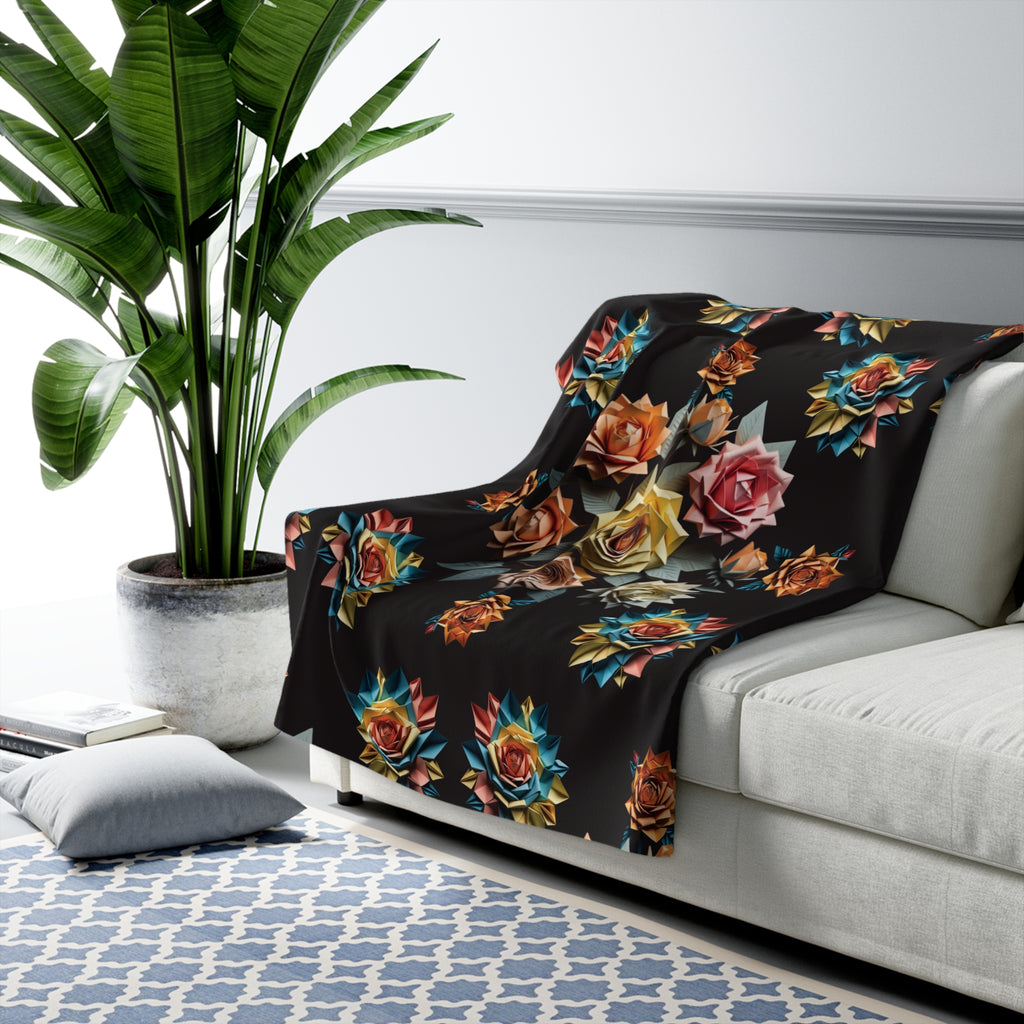 Black throw blanket with scattered geometric origami roses in amber, teal, gold, crimson, and pink tones with dark leaves draped over a sofa