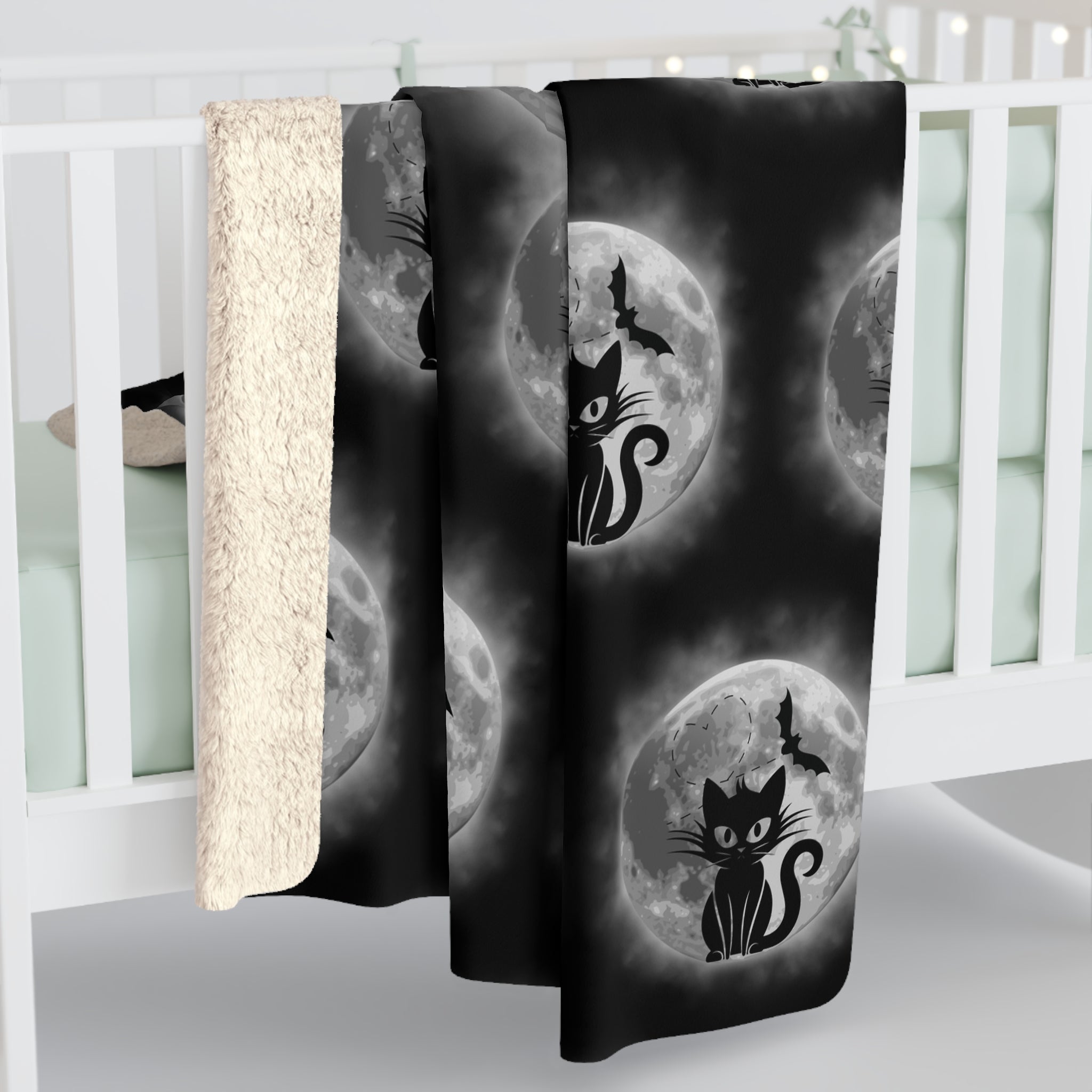 Black throw blanket with repeating pattern of black cat silhouettes seated before glowing full moons with bats in flight against a dark smoky background, shown draped over a white crib rail with sherpa back visible
