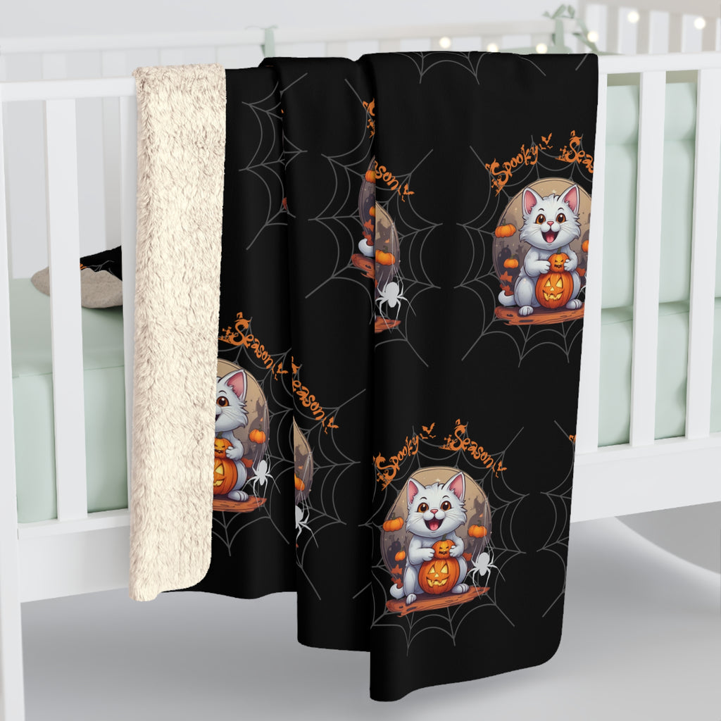 Black throw blanket with repeating pattern of a grinning white cat hugging a jack-o'-lantern beneath a full moon with mini pumpkins, dangling white spiders, and orange Spooky Season text over a grey cobweb background, shown draped over white crib rail with sherpa back visible