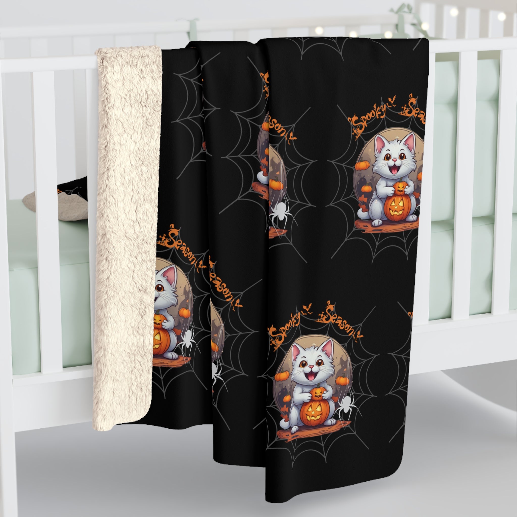 Black throw blanket with repeating pattern of a grinning white cat hugging a jack-o'-lantern beneath a full moon with mini pumpkins, dangling white spiders, and orange Spooky Season text over a grey cobweb background, shown draped over white crib rail with sherpa back visible