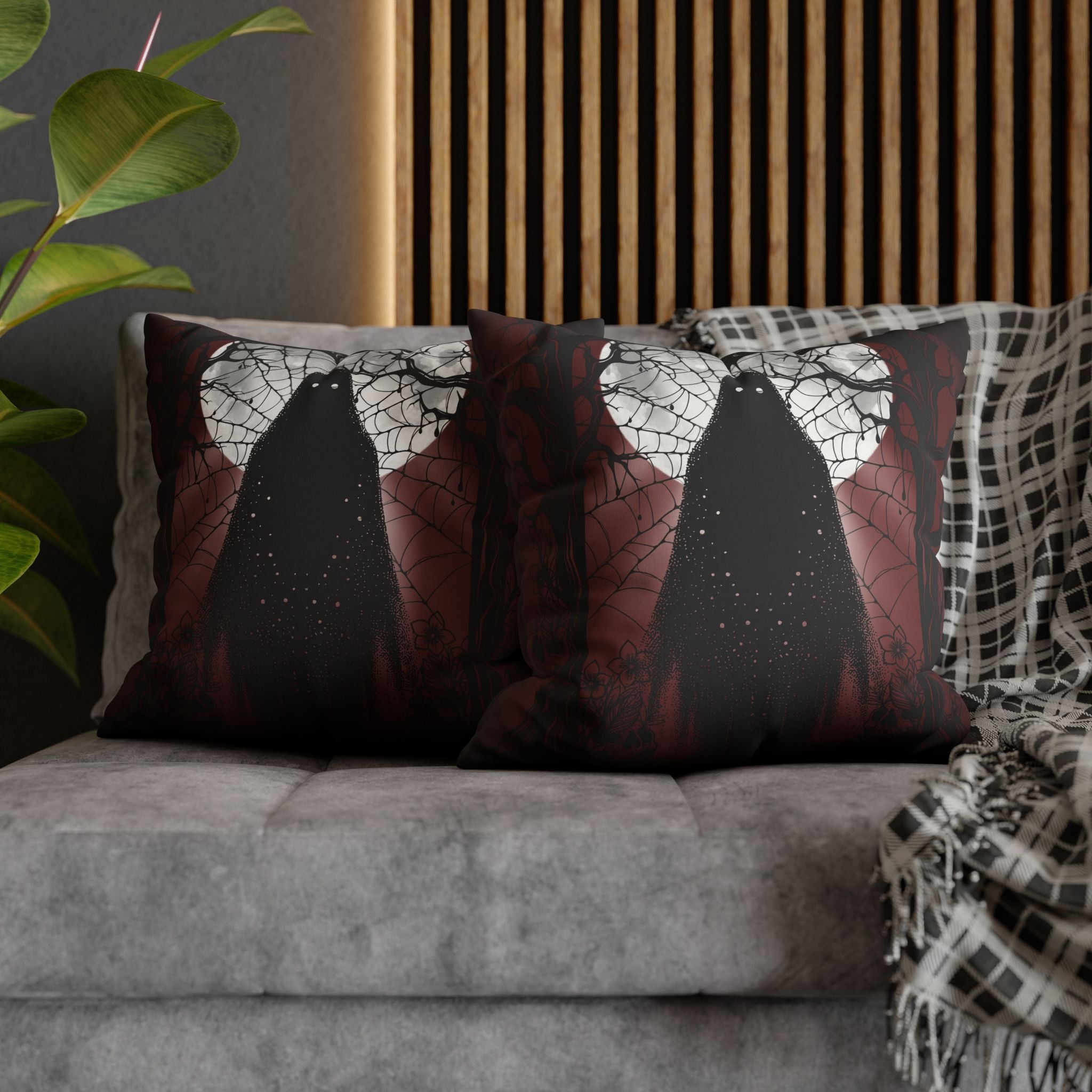 Two square throw pillows on a grey sofa featuring three tall dark ghost silhouettes with faint glowing eyes standing beneath a full moon wrapped in cobwebs and bare branching trees against a deep crimson bleeding into black background with white star-dust at their hems
