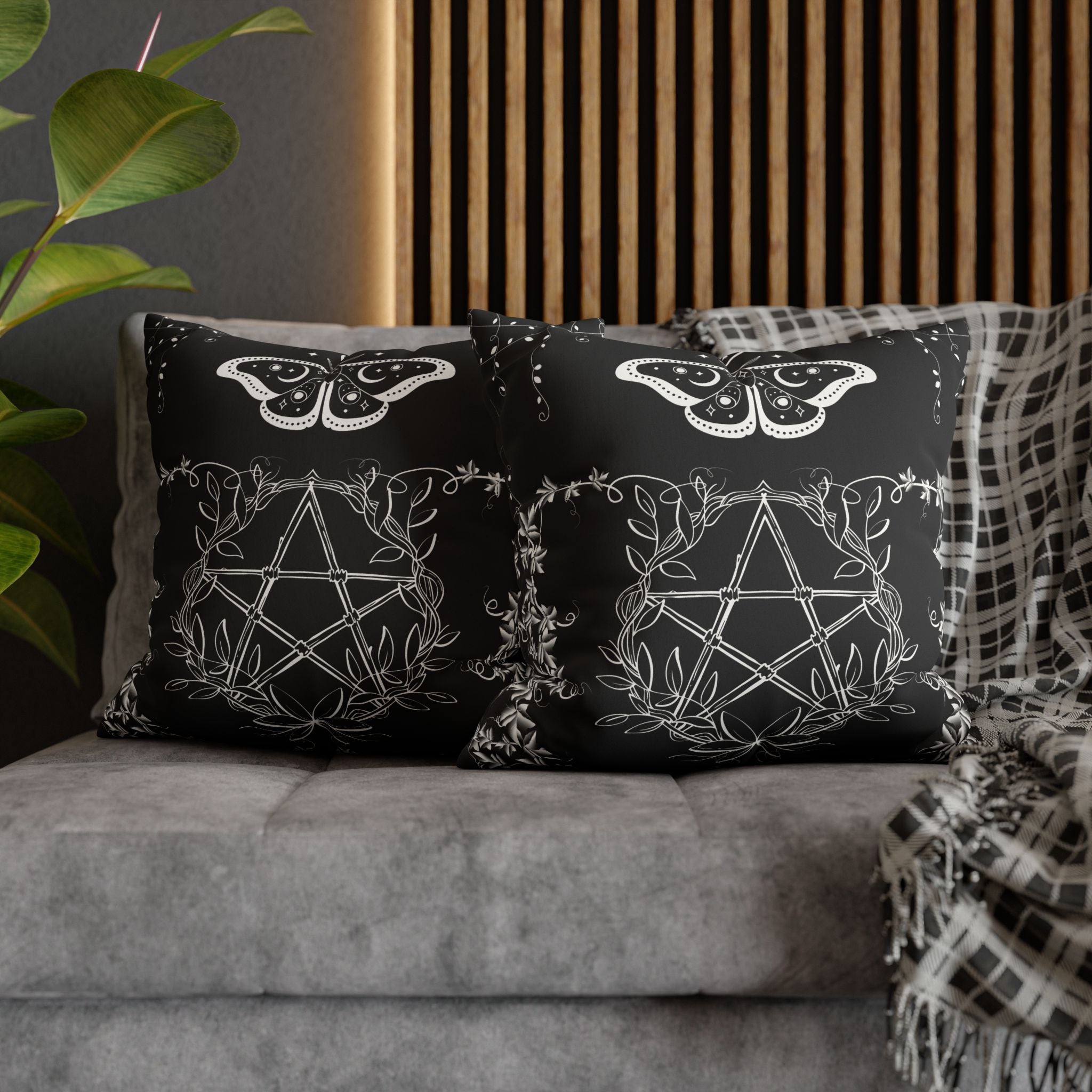 Two square throw pillows on a grey sofa featuring a white linework celestial moth with moon and star wing markings above a botanical pentagram woven with trailing vines and leaves against a deep black background