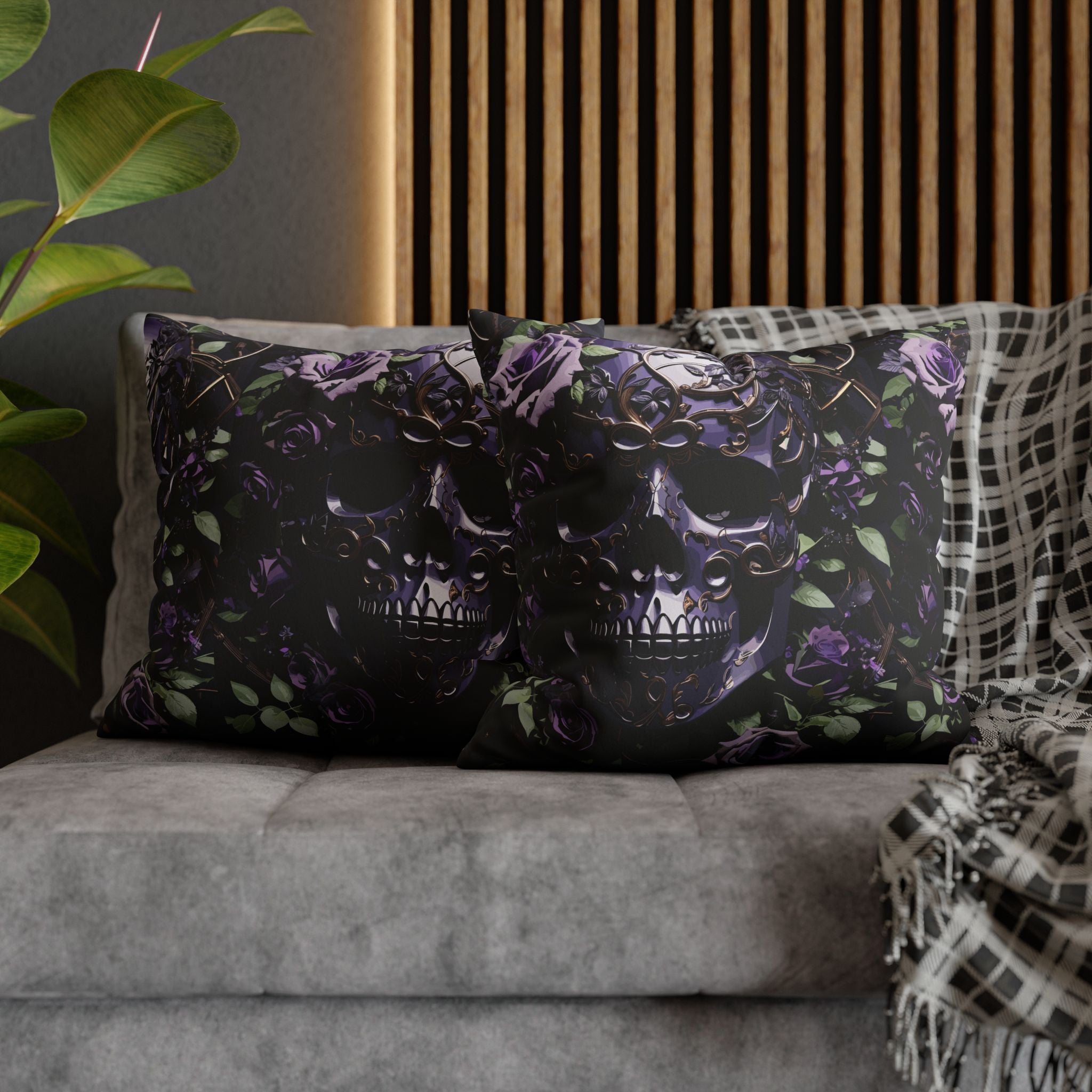 Two square throw pillows on a grey sofa featuring a large obsidian skull with hollow black eyes and purple and gold baroque filigree detailing surrounded by deep purple roses and green leaves against a black background