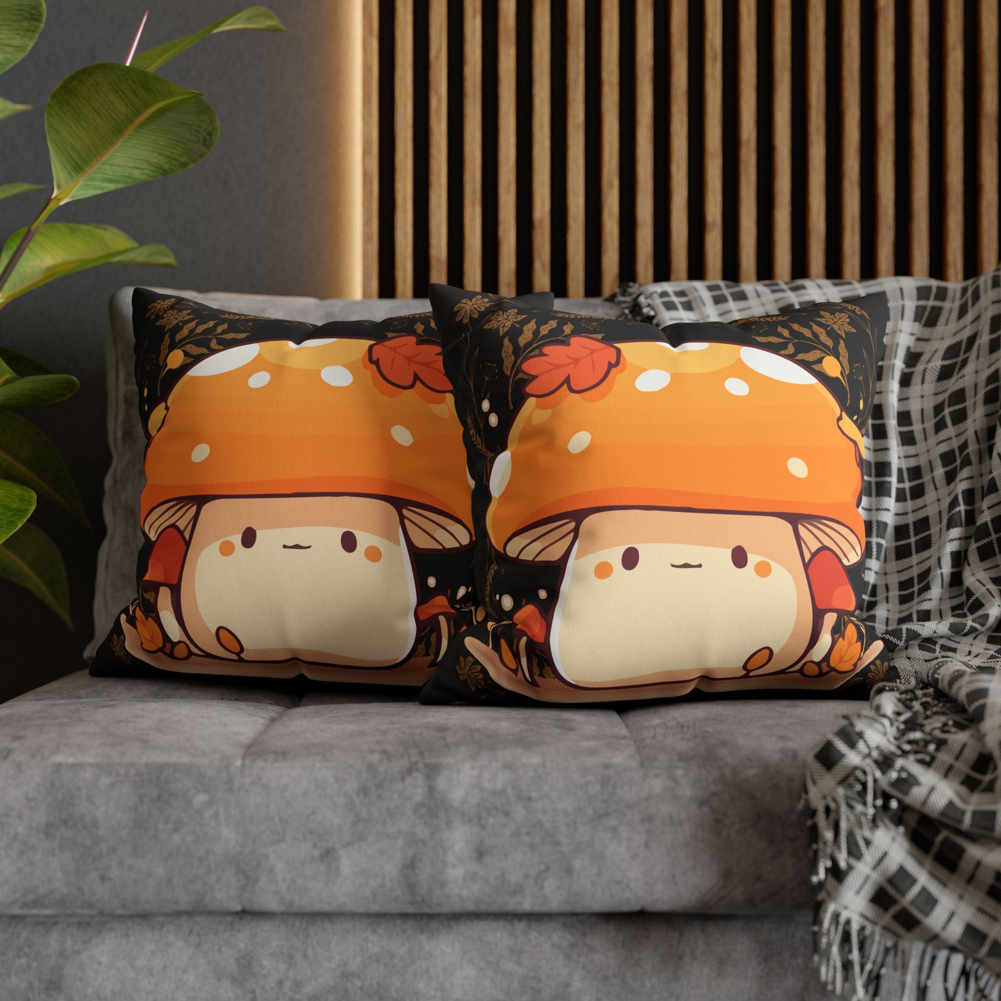 Two square throw pillows on a grey sofa featuring a round kawaii creature wearing a large orange and white spotted mushroom cap surrounded by tiny red mushrooms, autumn leaves, and golden botanical illustrations against a deep black background