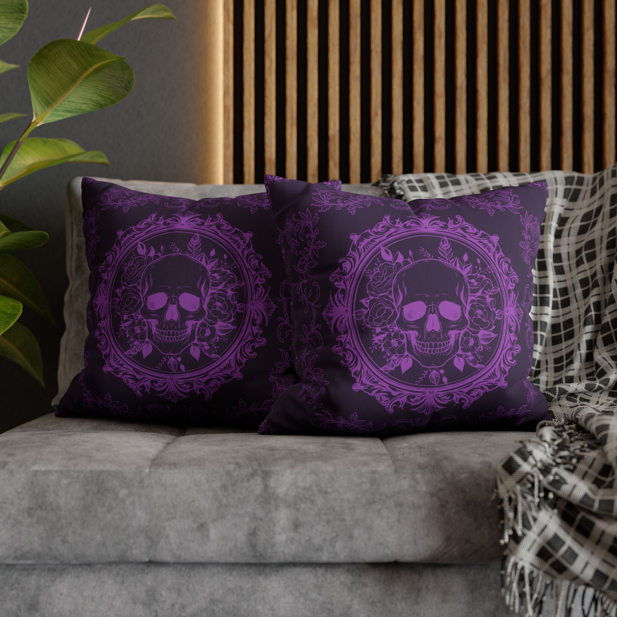 
Two square throw pillows on a grey sofa featuring a skull with flame-lit eyes inside an ornate circular baroque frame with roses and botanical flourishes and scattered filigree details rendered entirely in vivid violet purple on a deep plum black background