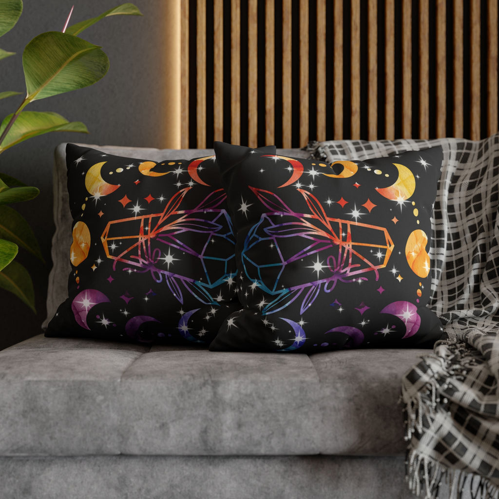 Two square throw pillows on a grey sofa featuring a symmetrical arrangement of rainbow gradient crystals in orange to purple tones with golden crescent moons above, deep purple crescents below, golden orbs, and white starburst sparkles scattered across a deep black background