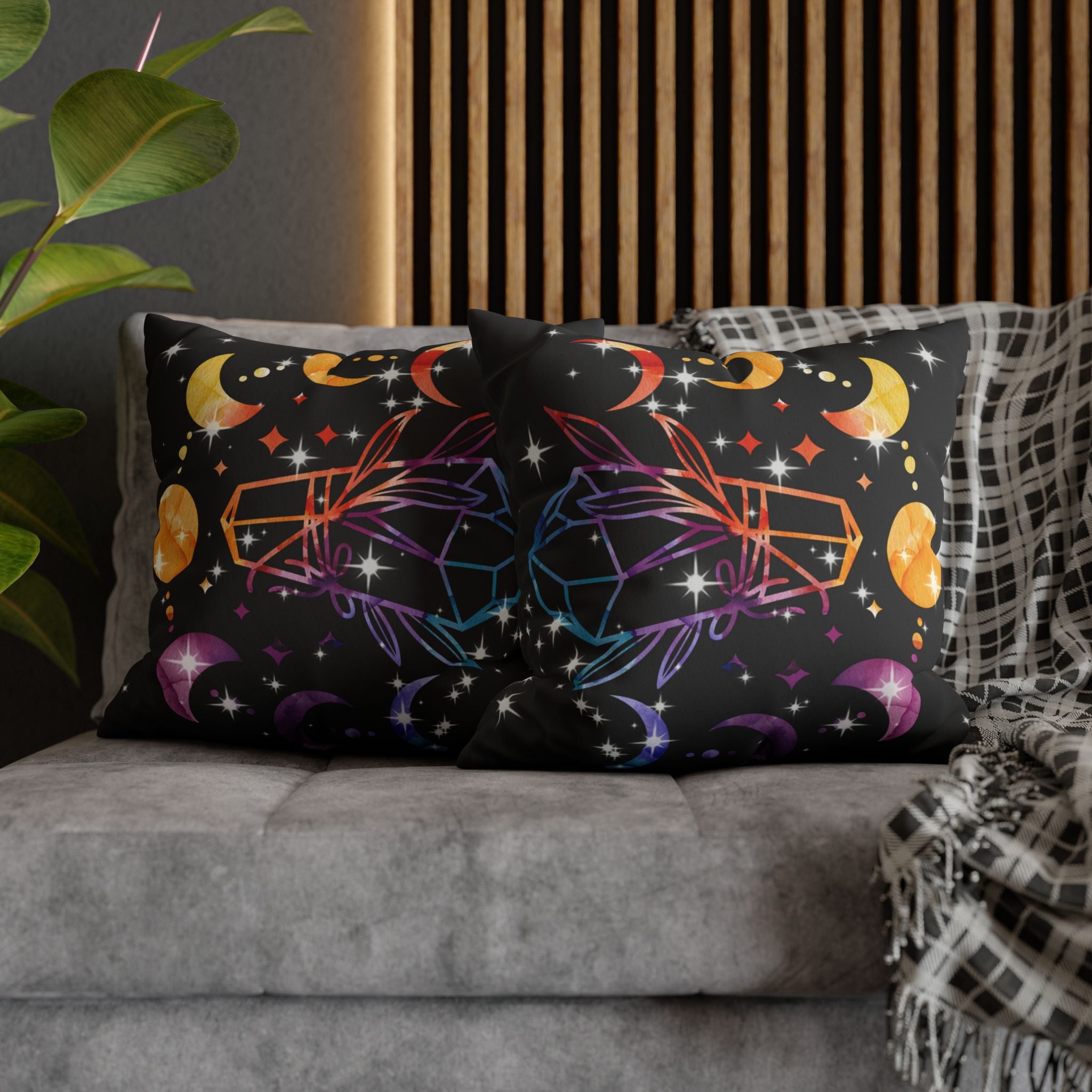 Two square throw pillows on a grey sofa featuring a symmetrical arrangement of rainbow gradient crystals in orange to purple tones with golden crescent moons above, deep purple crescents below, golden orbs, and white starburst sparkles scattered across a deep black background