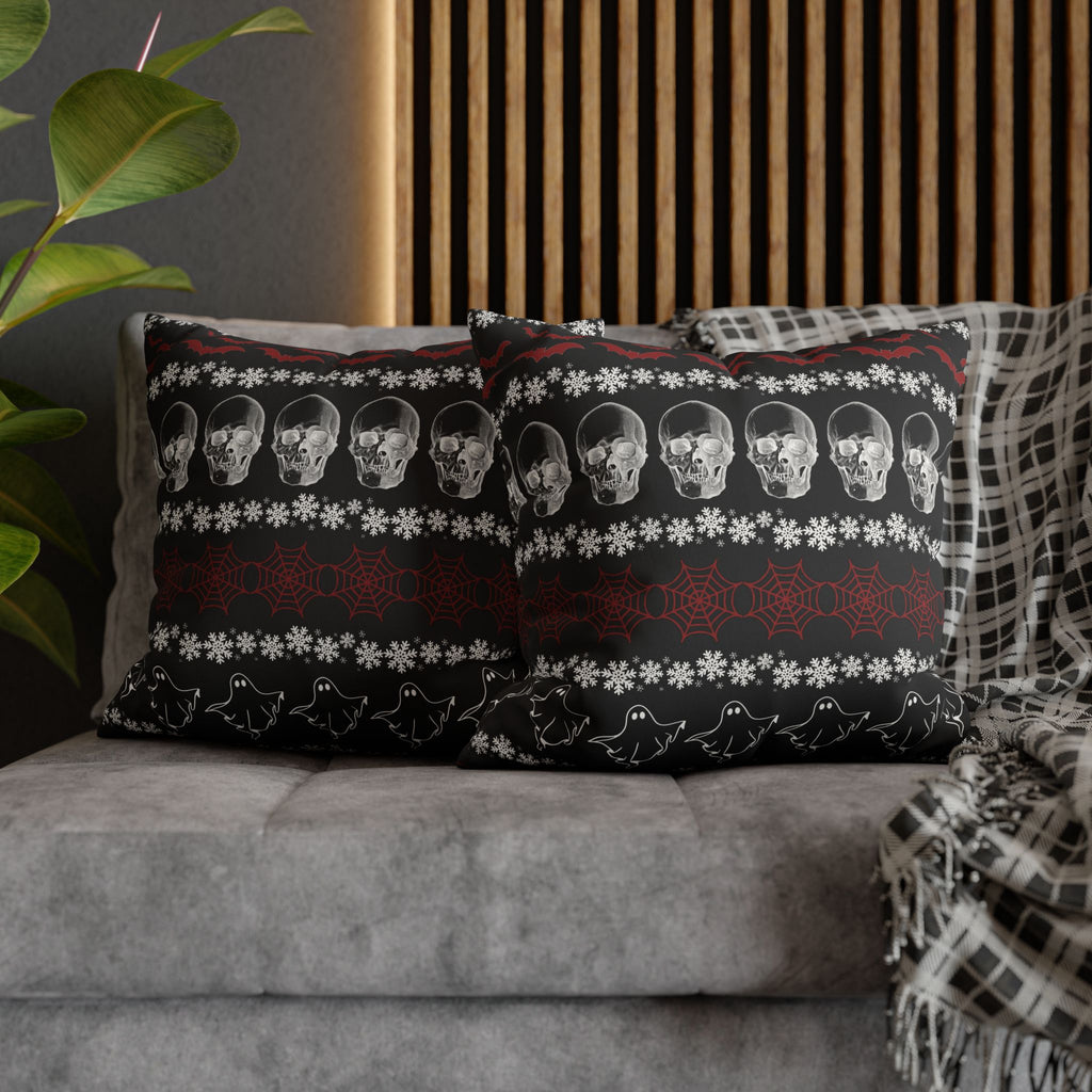Two square throw pillows on a grey sofa featuring a gothic ugly sweater pattern with horizontal stripes of grey skulls, red cobwebs, and white ghost outlines separated by white snowflake rows with red bats along the top on a deep black background