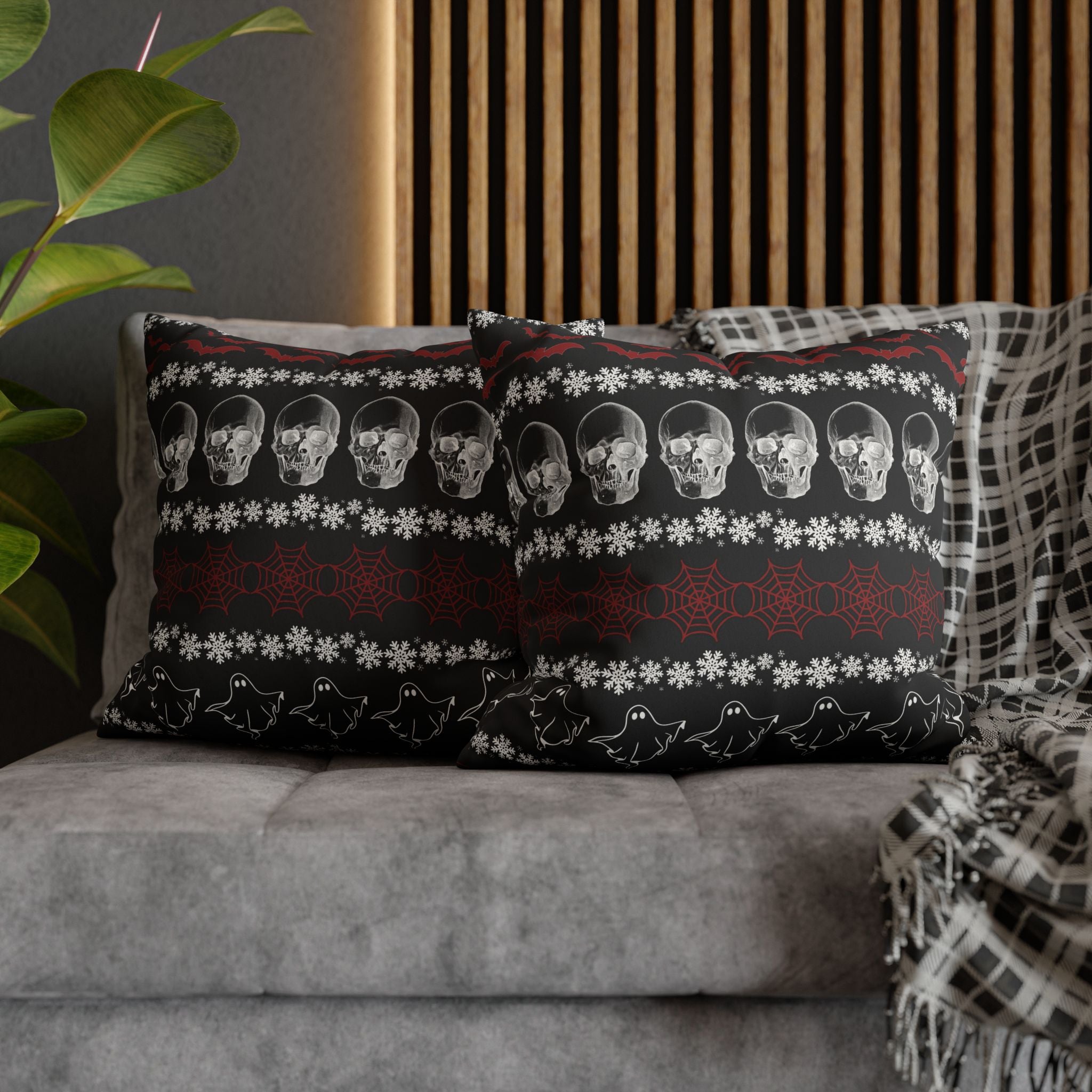 Two square throw pillows on a grey sofa featuring a gothic ugly sweater pattern with horizontal stripes of grey skulls, red cobwebs, and white ghost outlines separated by white snowflake rows with red bats along the top on a deep black background