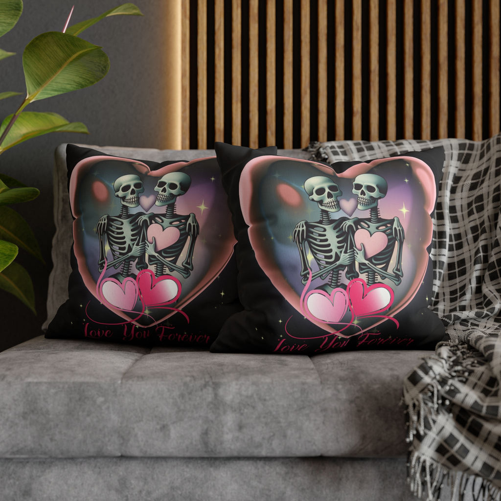 Two square throw pillows on a grey sofa featuring two skeletons leaning together inside a glowing pink heart frame with one holding a pink heart, a lavender heart floating between their skulls, pink ribbon hearts below, and Love You Forever in flowing pink script against a deep cosmic purple-teal backdrop on black