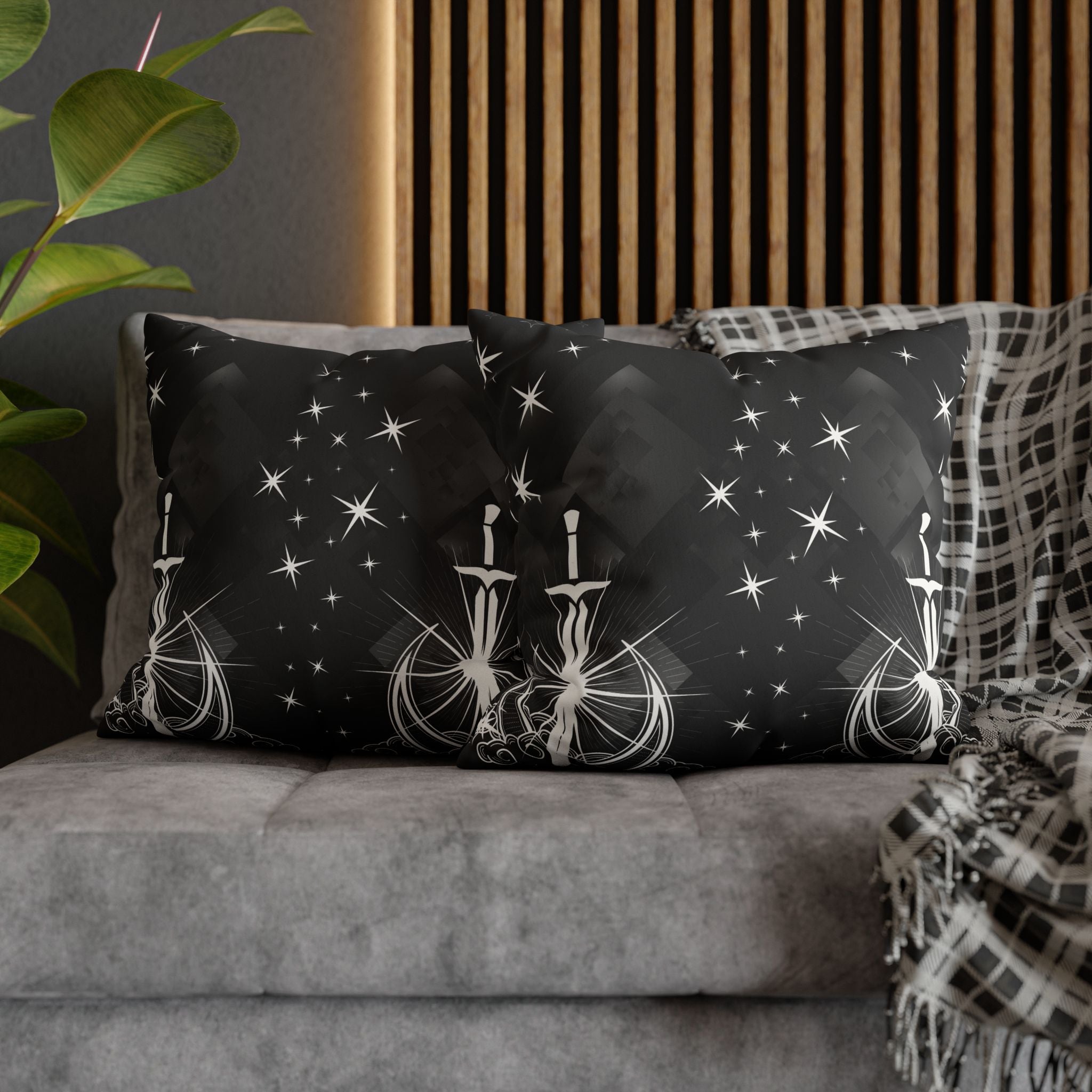 Two square throw pillows on a grey sofa featuring white linework swords piercing a crescent moon at the center of starburst rays rising from swirling clouds with scattered four and eight point stars and faceted geometric shapes against a deep black background