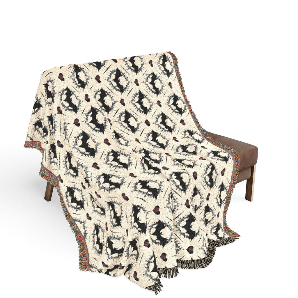 Woven cotton throw blanket with repeating bat silhouette and daisy pattern in cream and black with colorful fringed edges draped over a brown upholstered bench