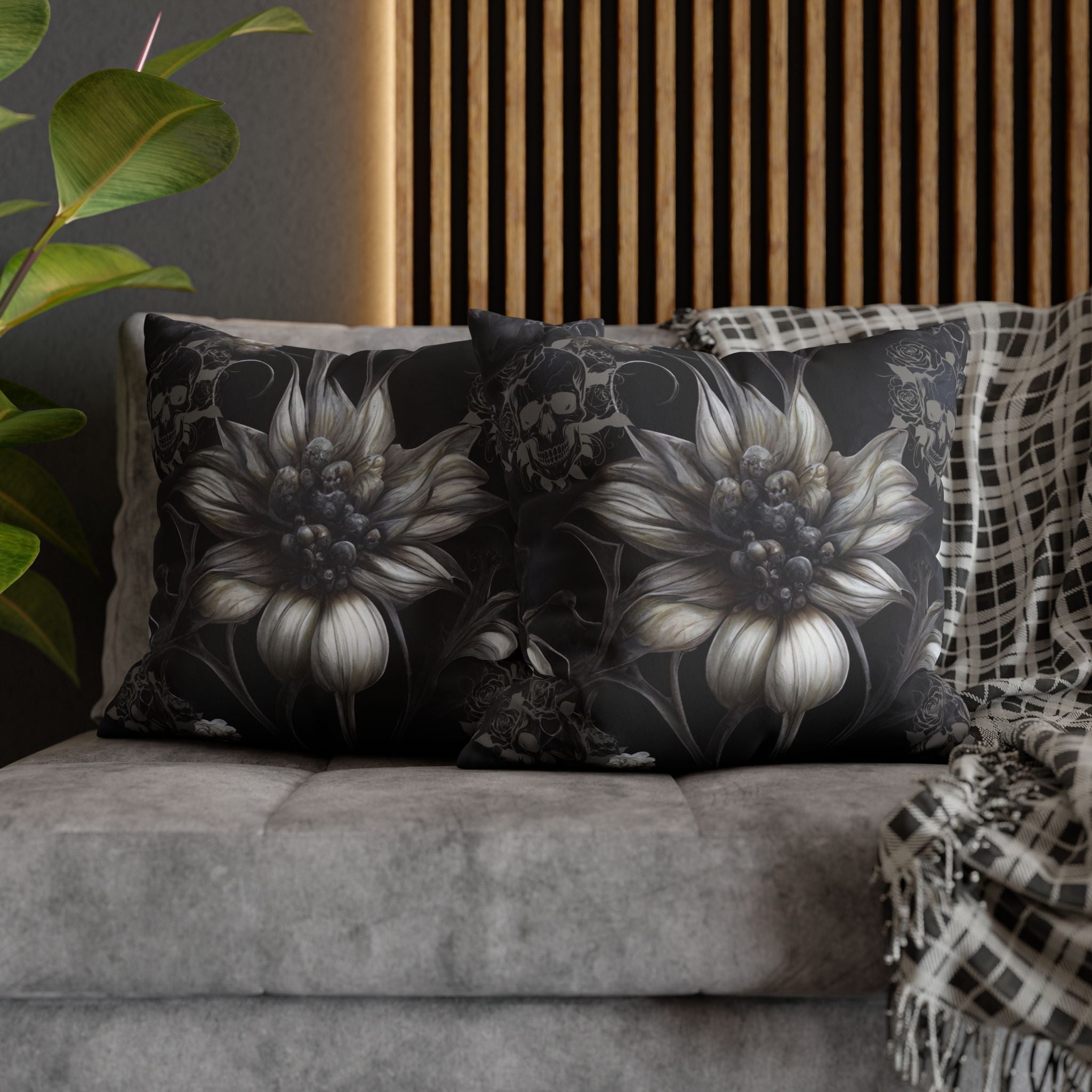 Two square throw pillows on a grey sofa featuring large ghostly white sunflowers with skull cluster centers in monochromatic grey and white with smaller skull and rose motifs drifting through the dark background connected by trailing vines against deep black