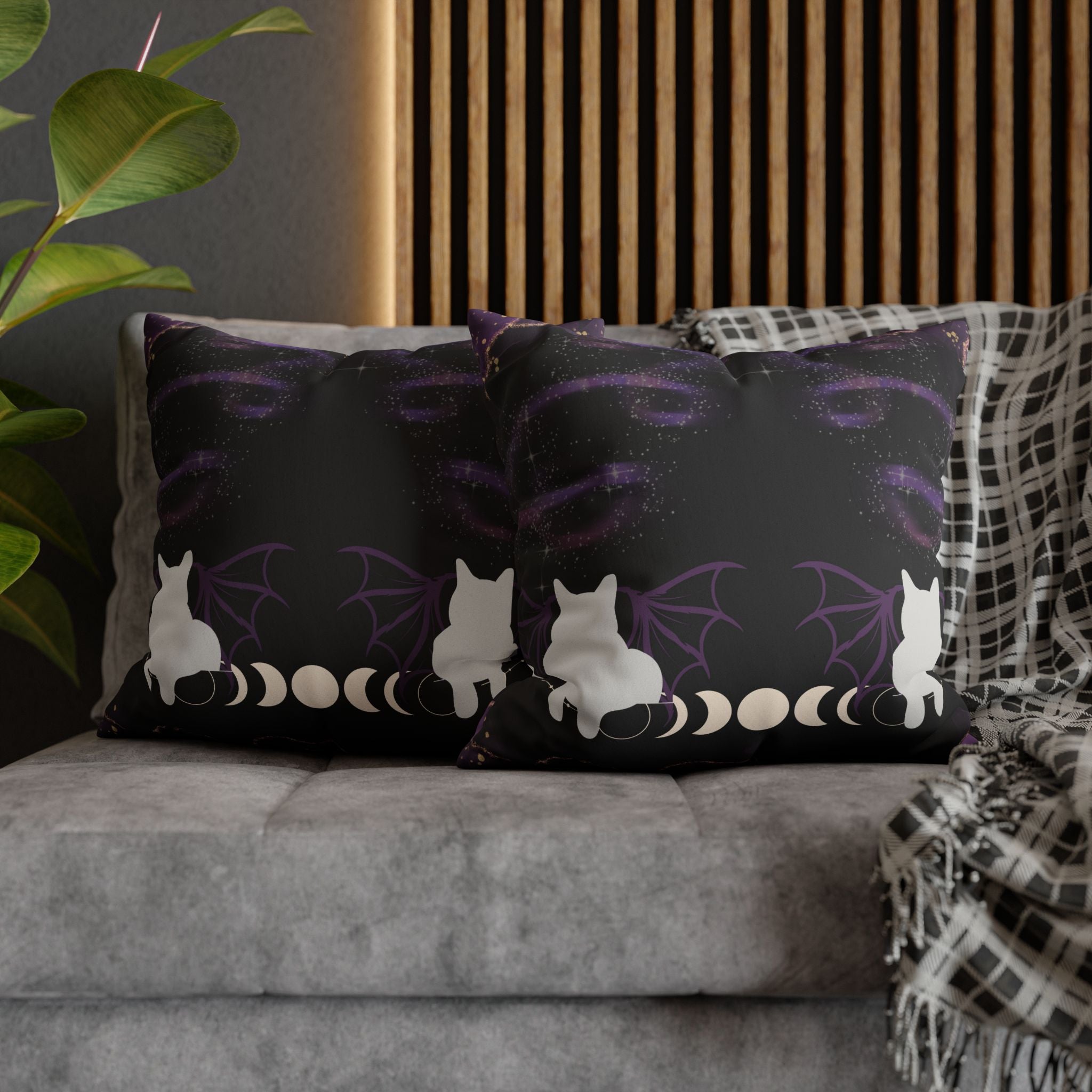Two square throw pillows on a grey sofa featuring a white cat silhouette viewed from behind with large dark bat wings spread wide against a swirling purple galaxy sky with the full moon phase sequence lined up along the bottom on deep black