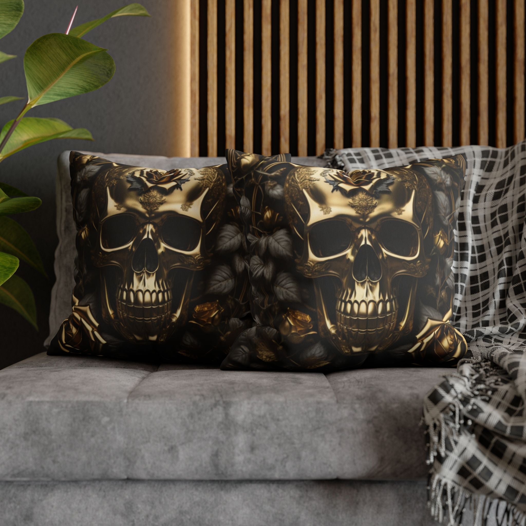 Two square throw pillows on a grey sofa featuring a burnished gold skull with ornate filigree forehead detailing surrounded by nearly-black roses and deep charcoal botanical leaves against a dark gold and black background