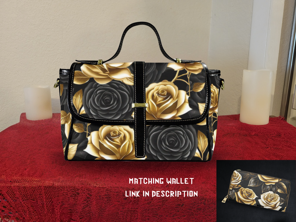 Black canvas satchel bag covered in gold and black rose floral print, black vegan leather trim, gold magnetic closure tab, brown adjustable shoulder strap, sitting on red tablecloth with candles