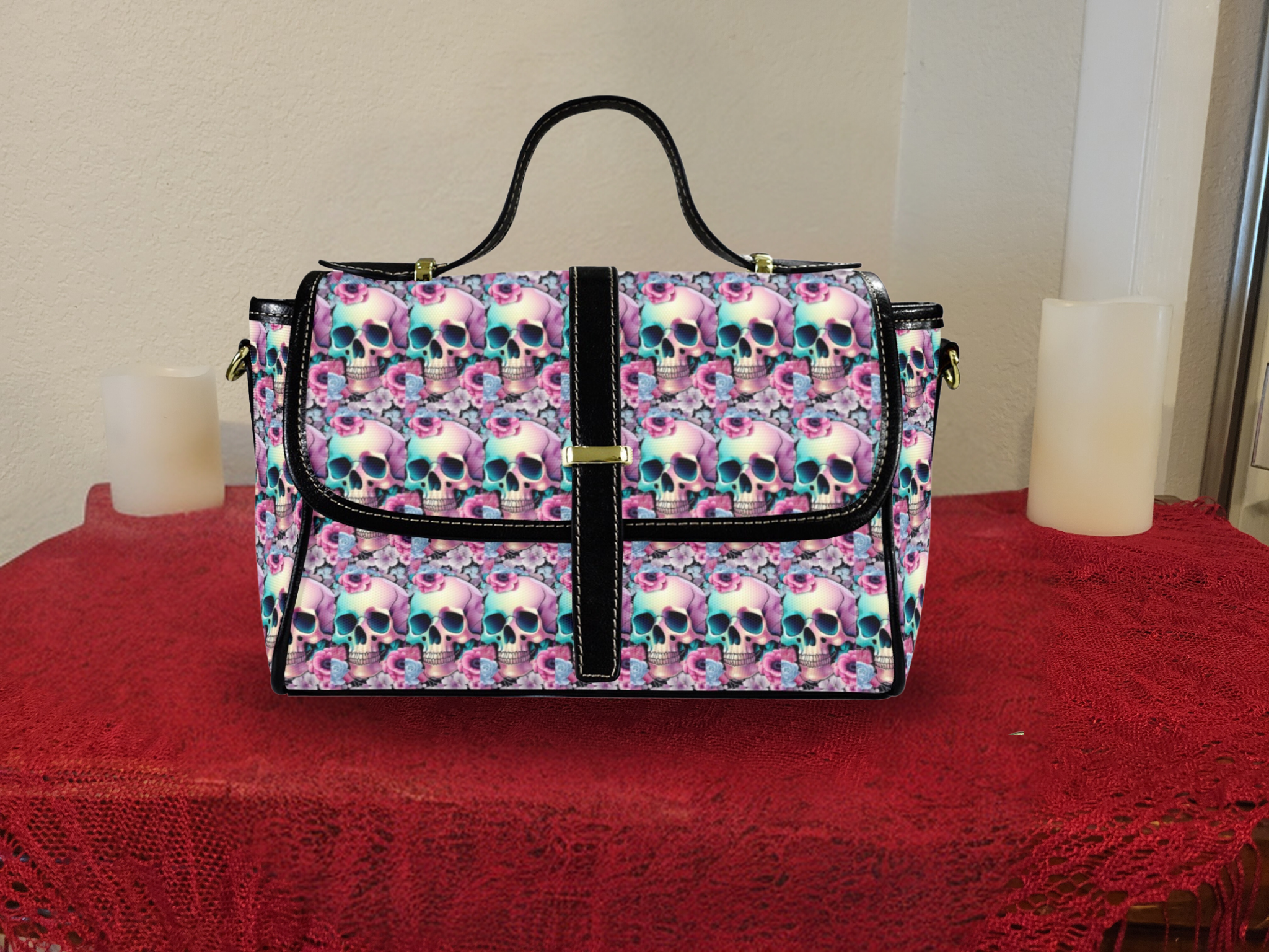 Black canvas satchel bag covered in repeating pattern of pastel gradient skulls and pink purple florals, black vegan leather trim, gold magnetic closure tab, sitting on red tablecloth with candles