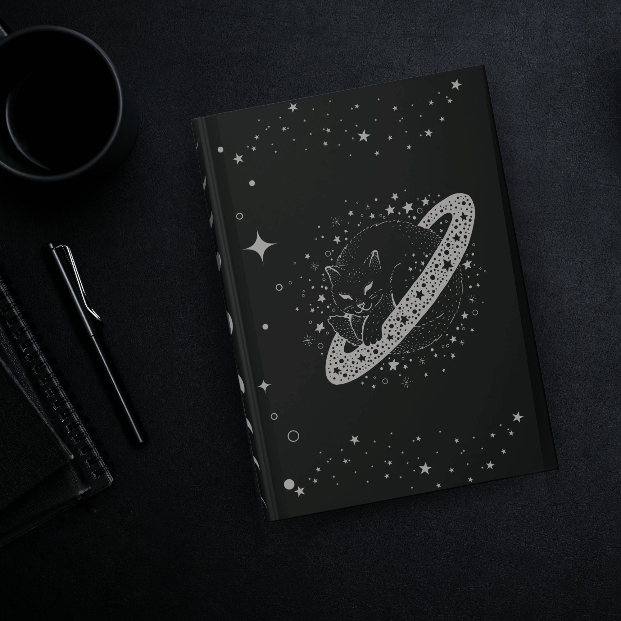 
Gothic hardcover journal with grey linework black cat curled inside a Saturn-like planetary ring surrounded by stars and cosmic dots on deep black background